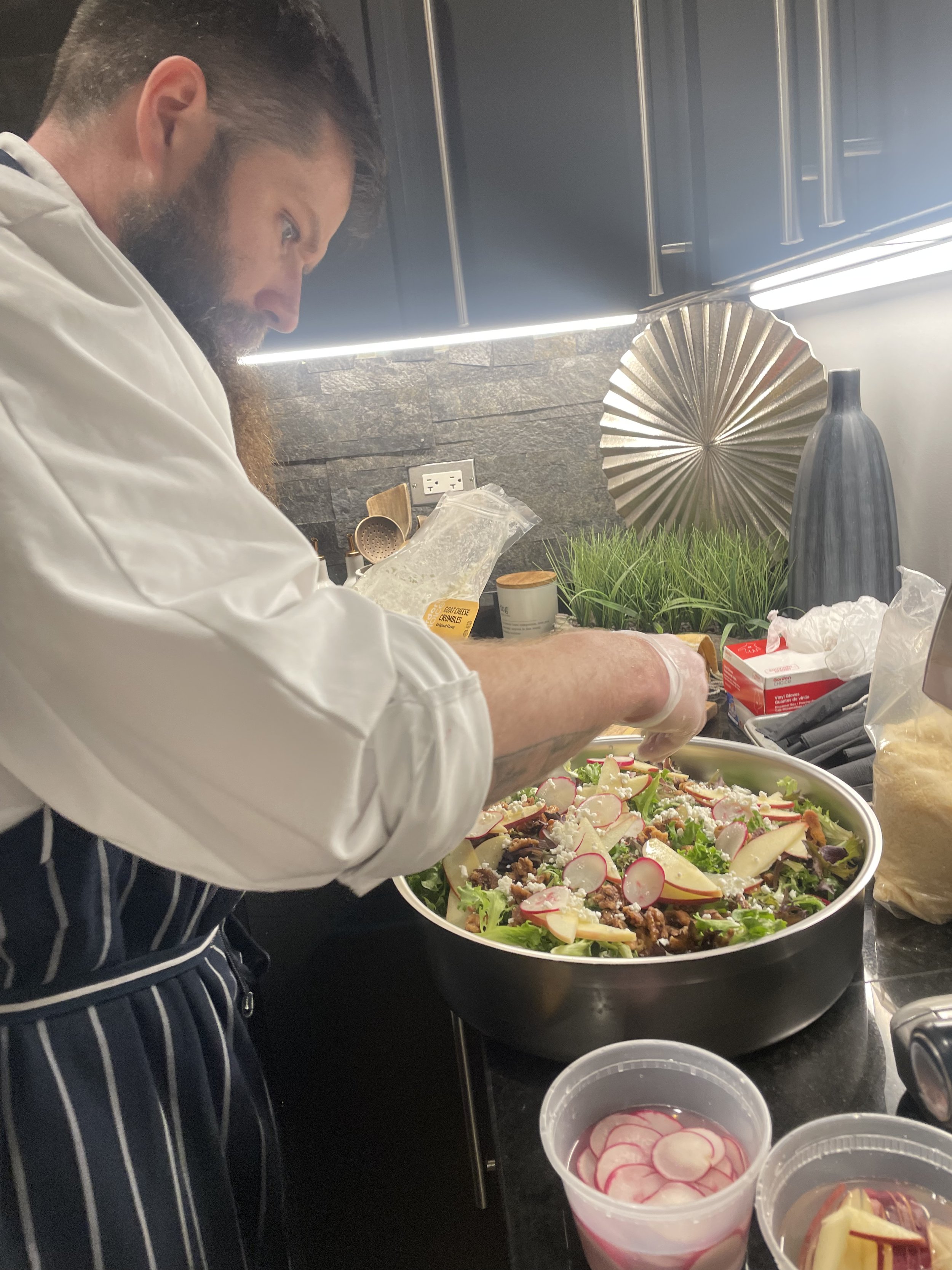 A man with a beard and dark hair preparing a large salad in a commercial kitchen. The salad contains radishes, cheese, and greens. The man is wearing a white chef's coat and a striped apron. Various kitchen utensils and ingredients are on the counter, and there is a metallic decorative piece and a power outlet on the wall behind him.