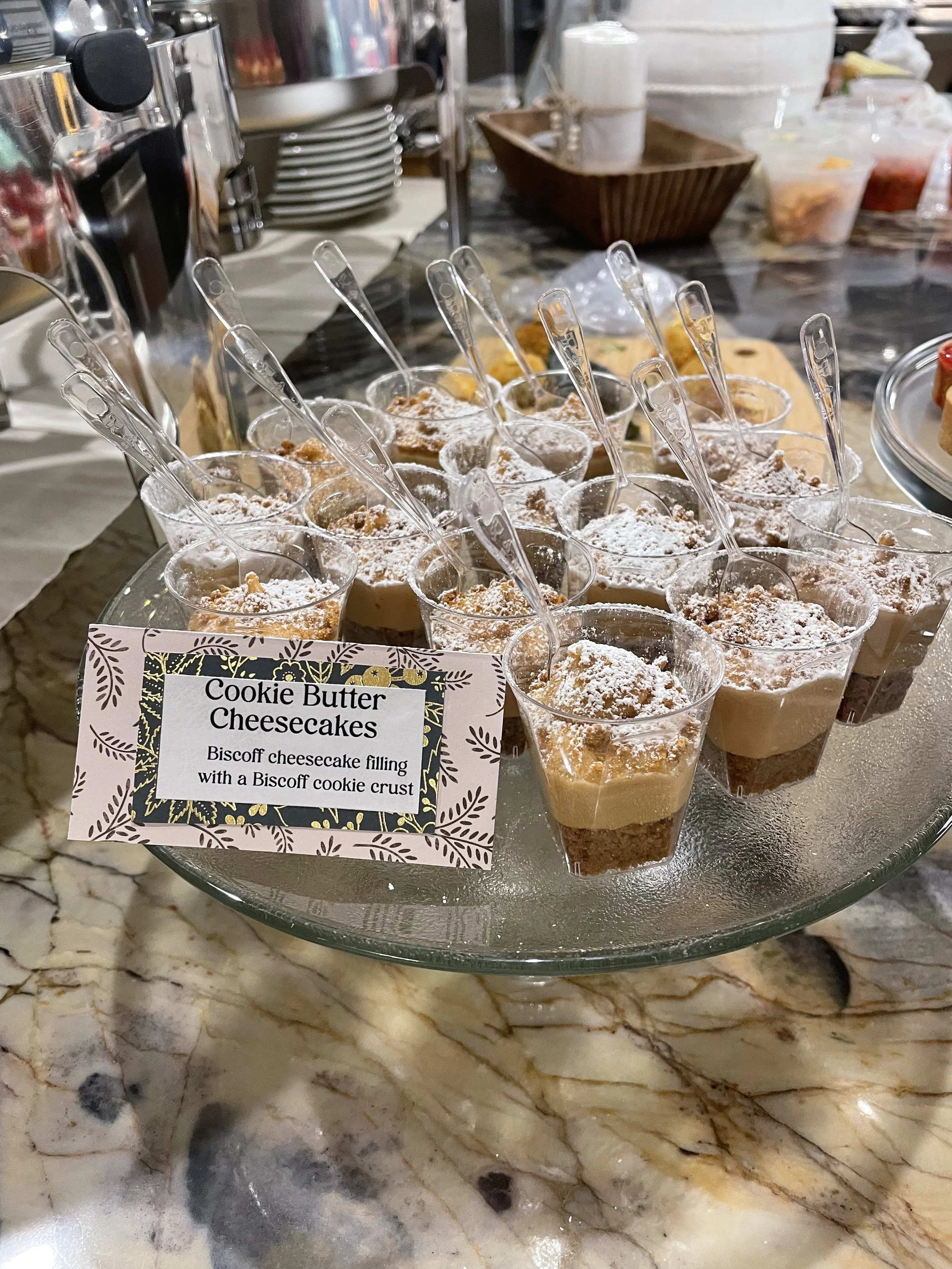 Plastic cups filled with cookie butter cheesecake, topped with powdered sugar and cookie crumbs, with small clear spoons inside each cup. A sign in front describes the dessert as 'Cookie Butter Cheesecakes' with a biscuit cookie crust.