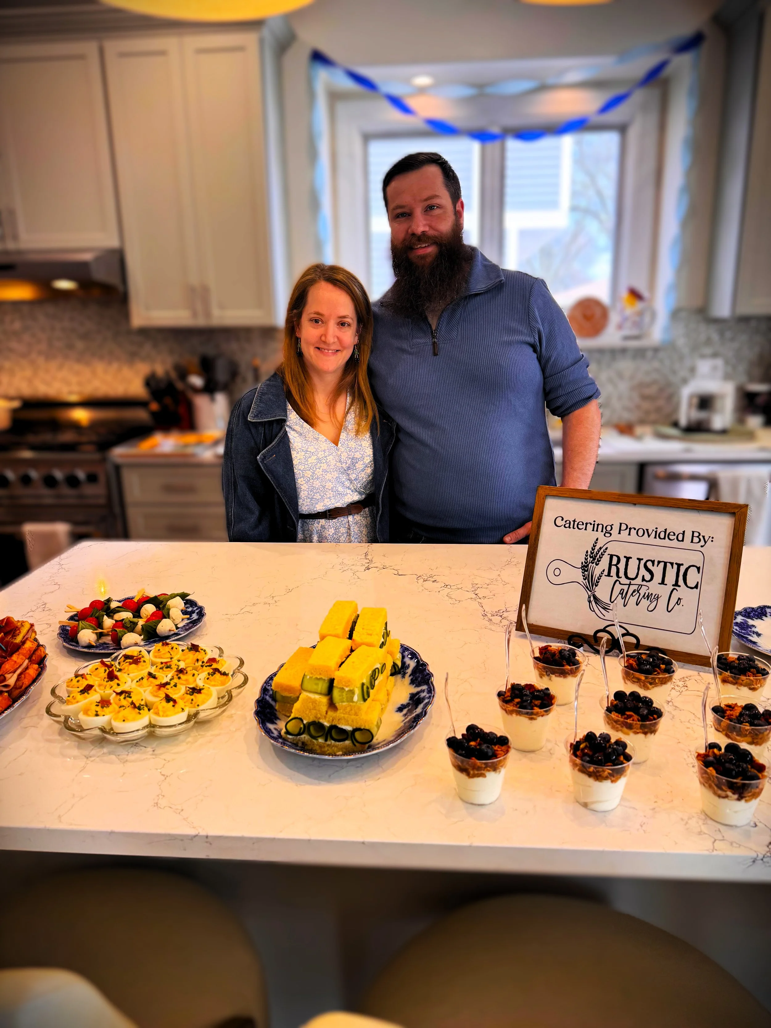 A woman and a man standing behind a table with assorted desserts, including mini cheesecakes, fruit-topped pastries, and layered desserts in clear cups, at a catered event. The table has a sign that reads 'Catering Provided By: Rustic Catering Co'.