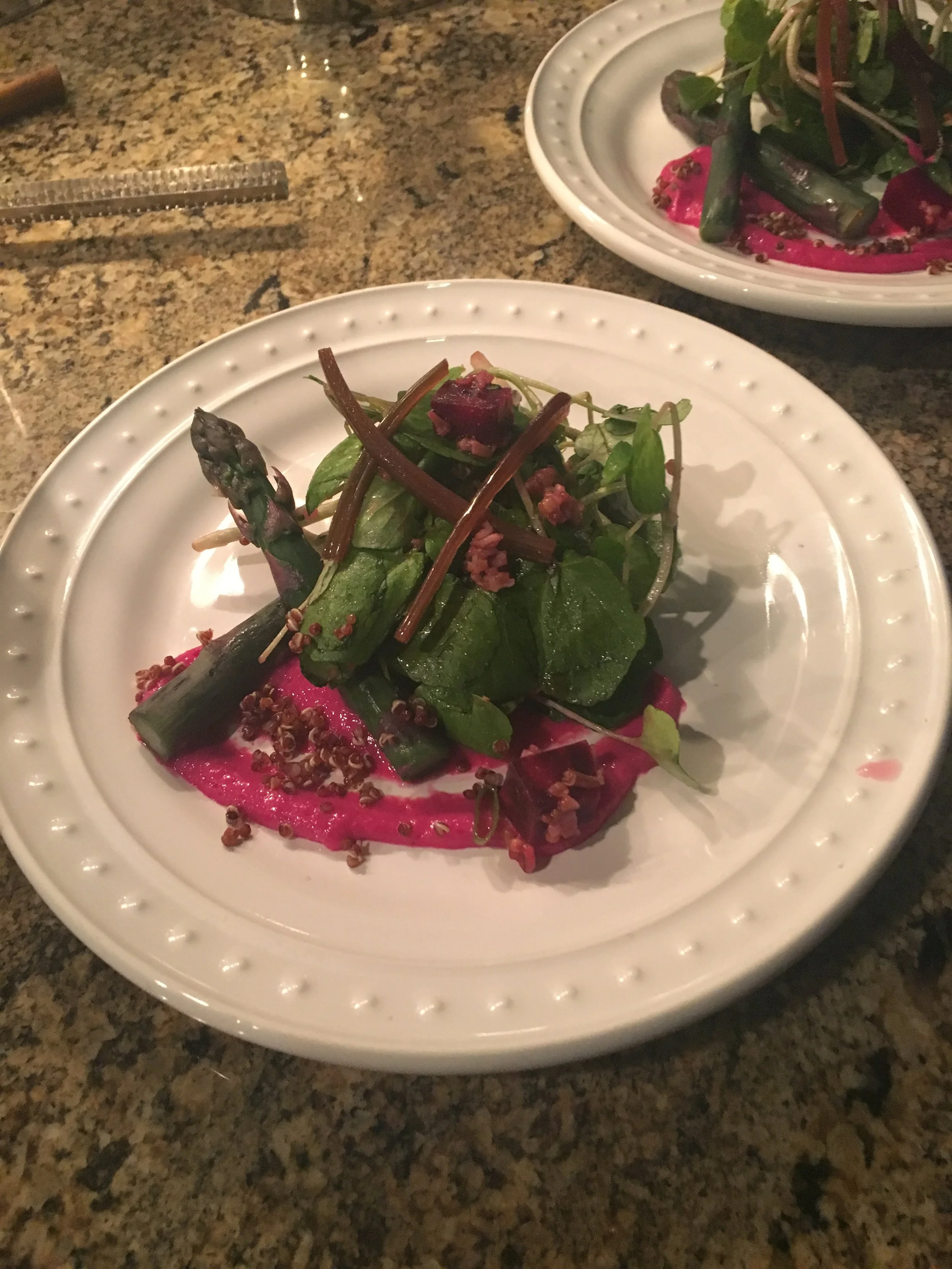 A plate with a salad including asparagus, spinach, and a pink sauce, topped with small seeds or grains, on a granite countertop.