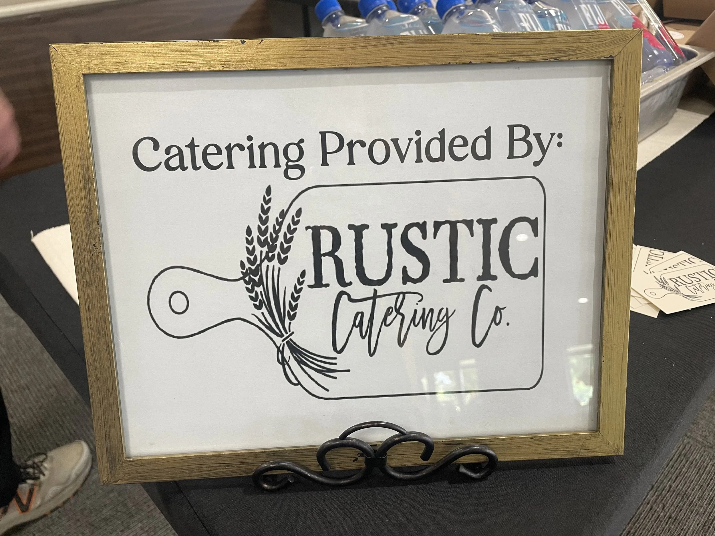 A sign with a wooden border that reads 'Catering Provided By: RUSTIC Catering Co.' with a decorative wheat illustration. In the background, bottled water and paper items are visible.