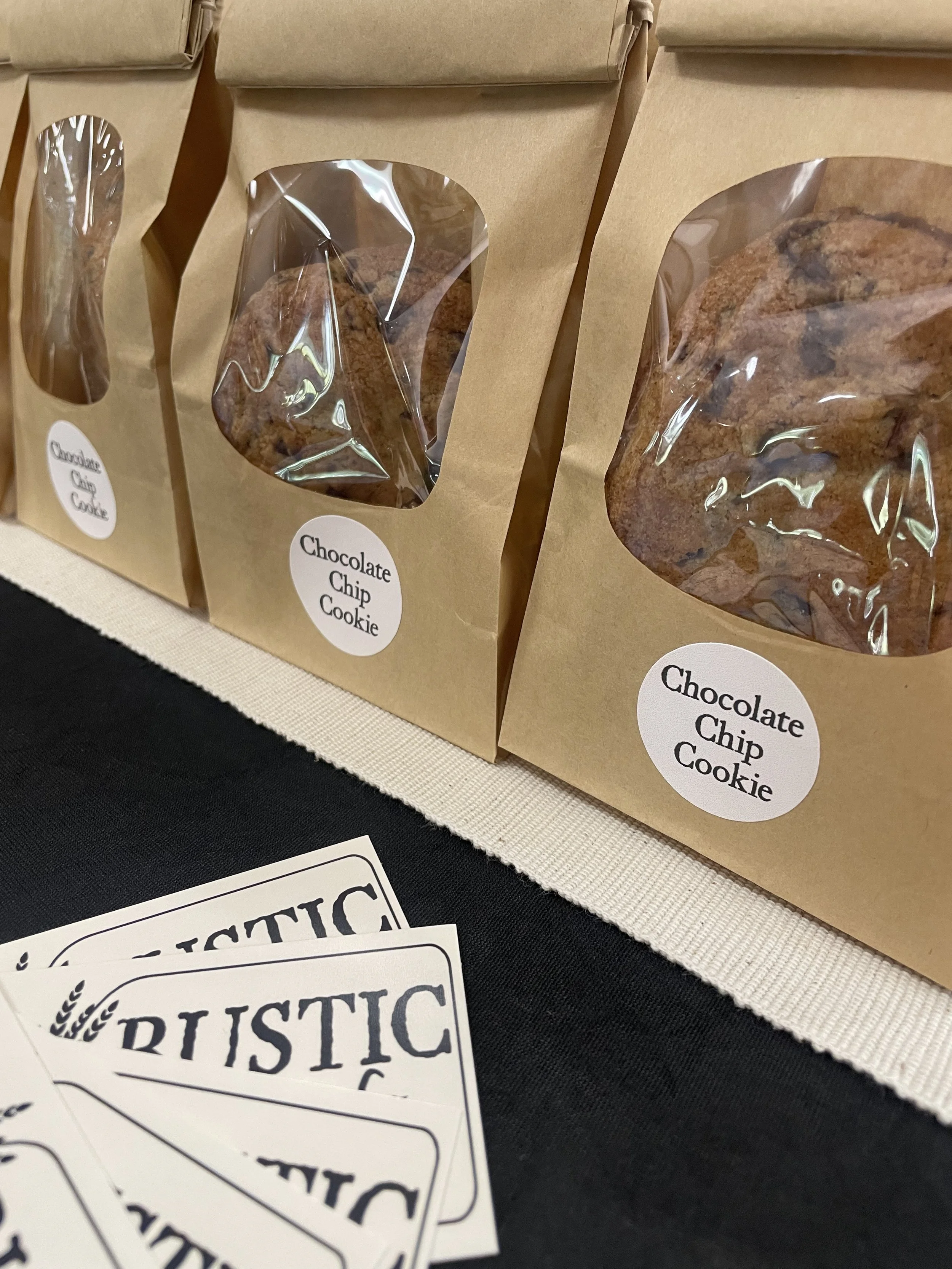 A display of brown paper bags with clear windows labeled "Chocolate Chip Cookie" on a table with black and white paper signs.