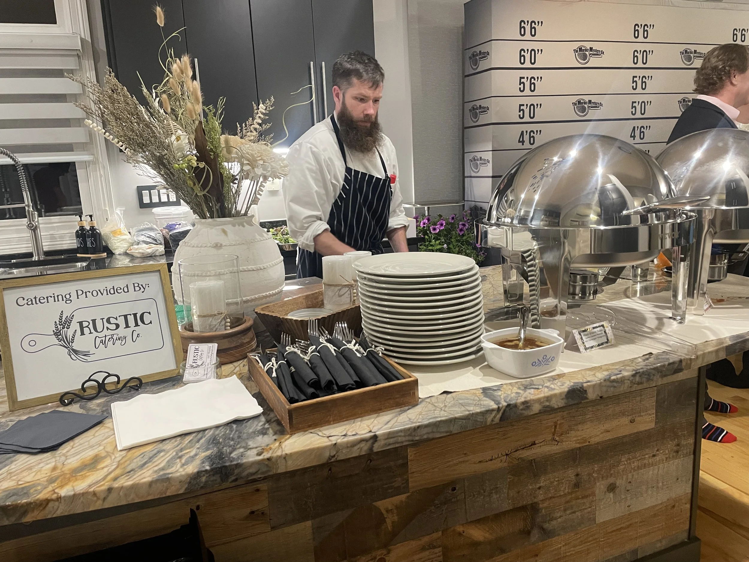 A man with a beard and striped apron standing behind a counter with plates, utensils, and a bowl of dipping sauce, in a room with a height measurement chart on the wall and a large pitcher with dried flowers nearby.
