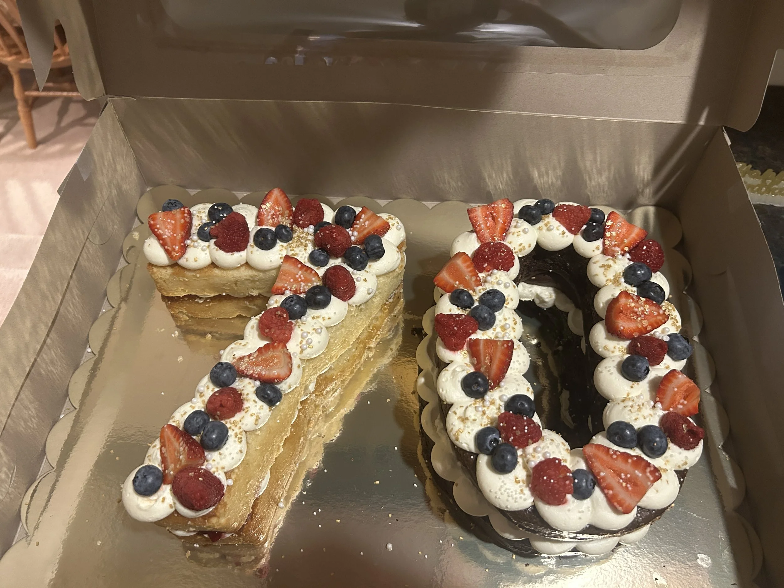 A partially sliced cake in a box topped with whipped cream and fresh berries, including strawberries, blueberries, and raspberries.