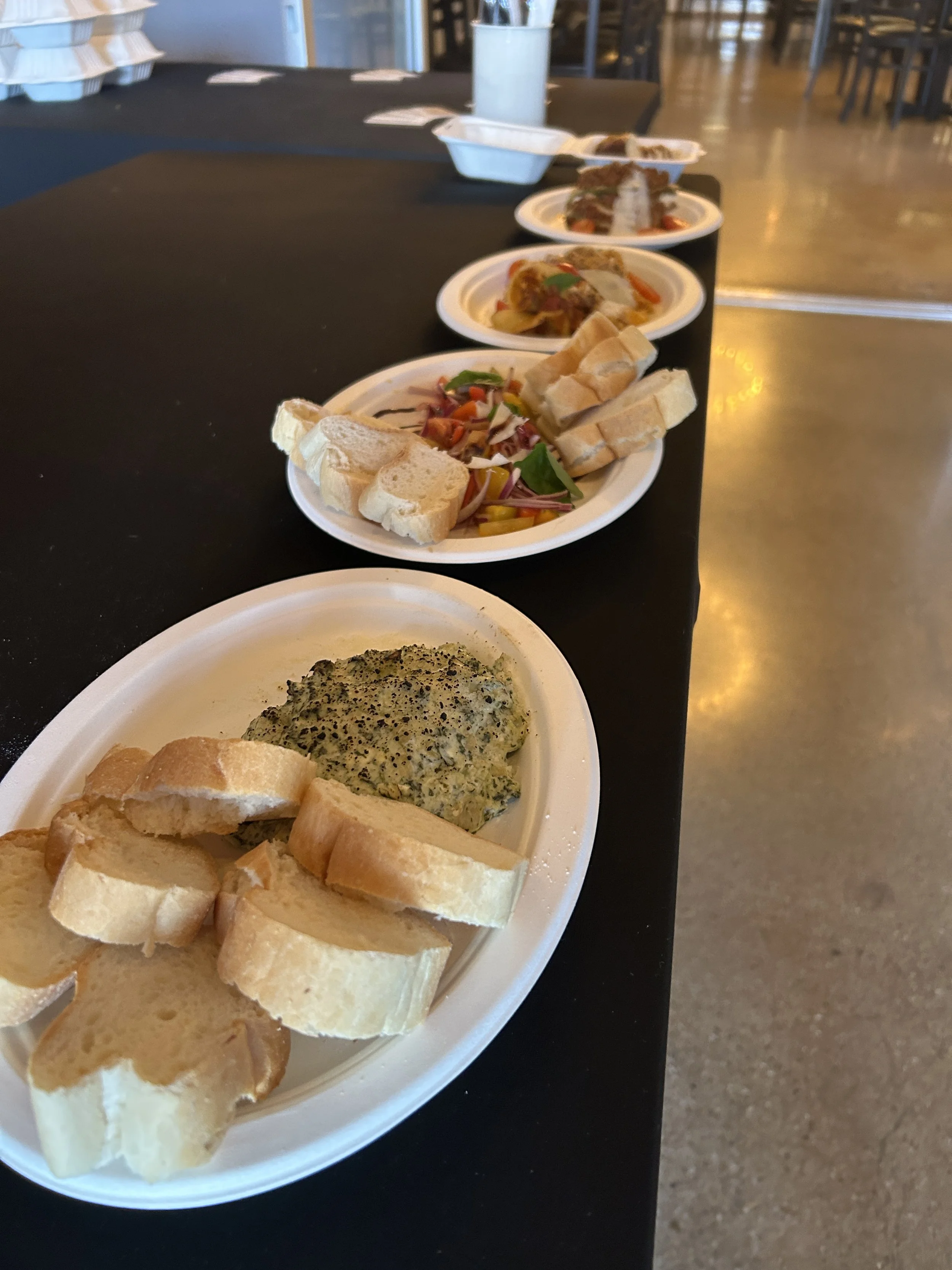 Plates with slices of bread, a spinach dip, and assorted salads on a black table in a cafeteria or dining area.