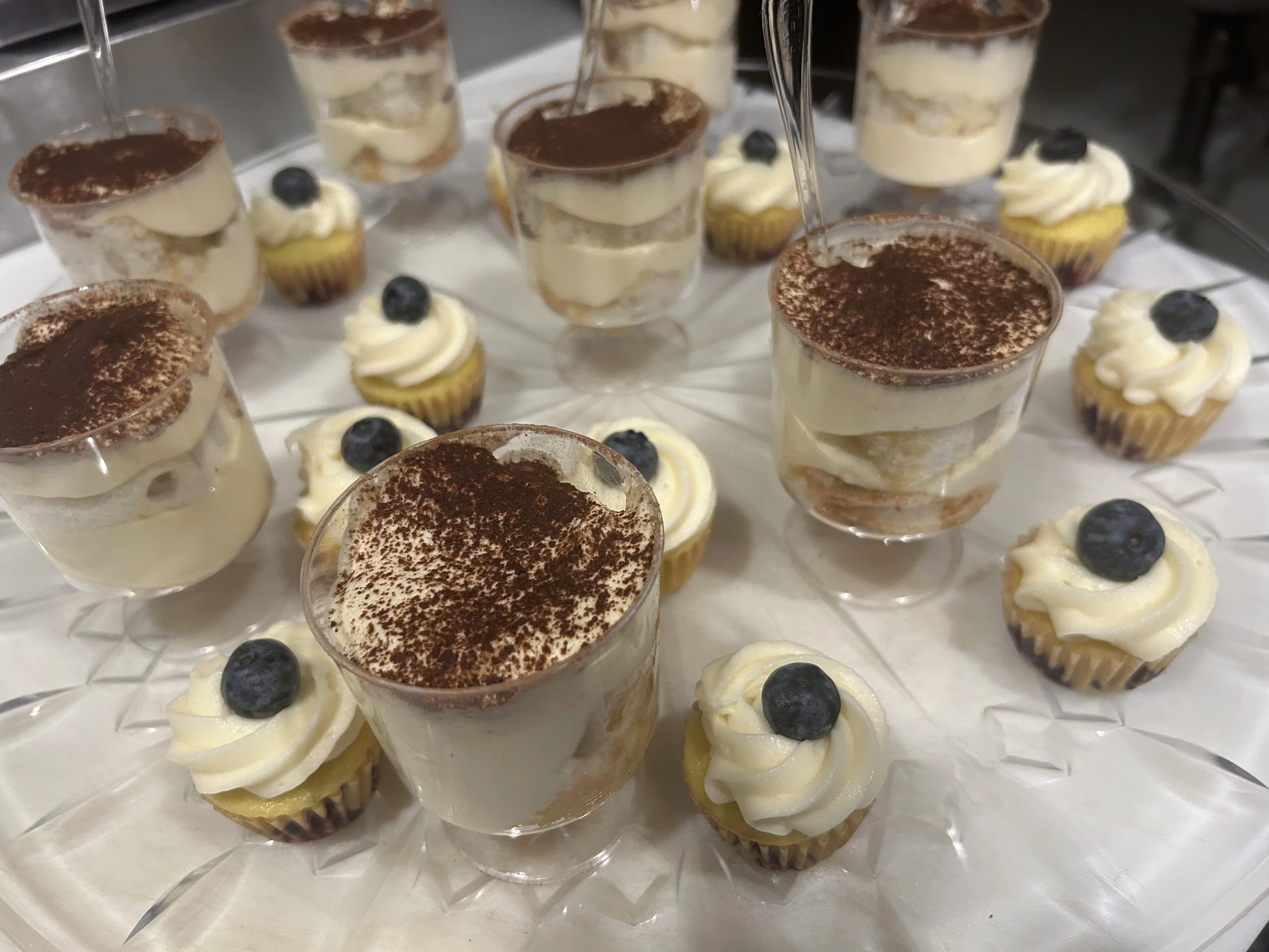 A tray of individual desserts in clear glasses and small cupcakes with blueberries on top. The desserts appear to be layered with cream or custard and topped with cocoa powder.