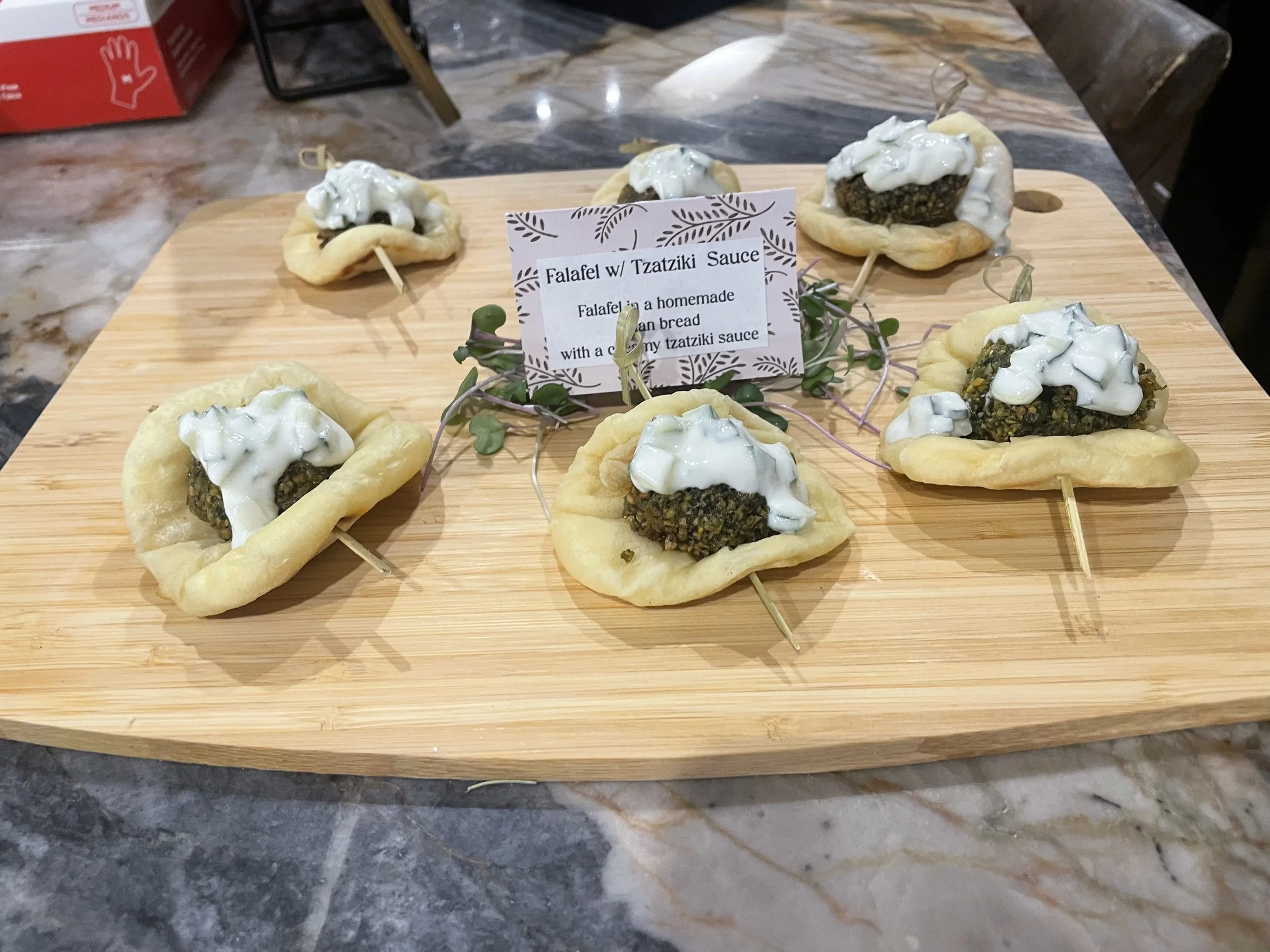 Several mini falafel sandwiches with creamy tzatziki sauce on a wooden serving board, garnished with small green plant leaves.