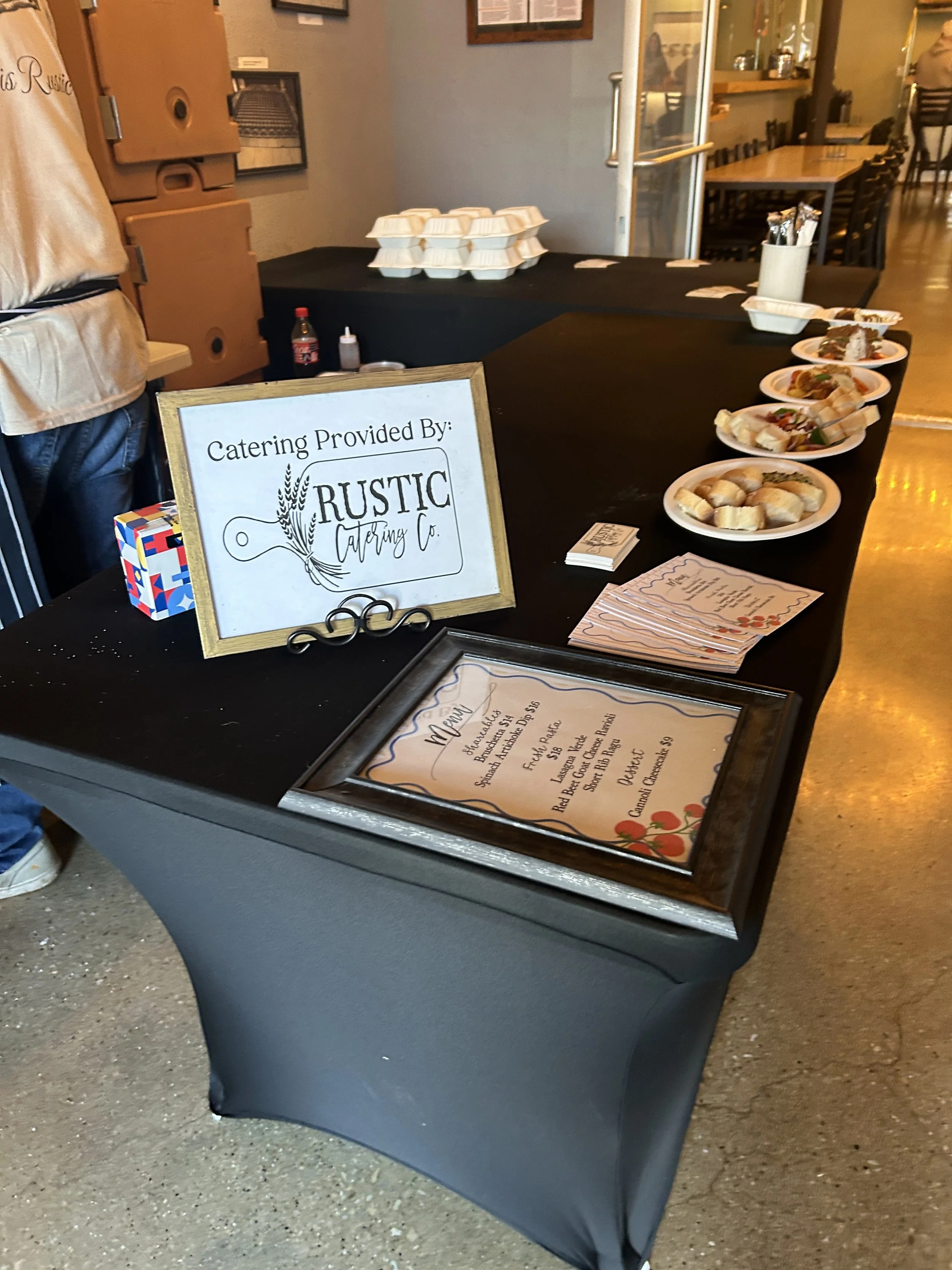 A catering table at a restaurant with plates of food, a framed menu, and a sign that reads 'Catering Provided By: Rustic Catering Co.' in the foreground.