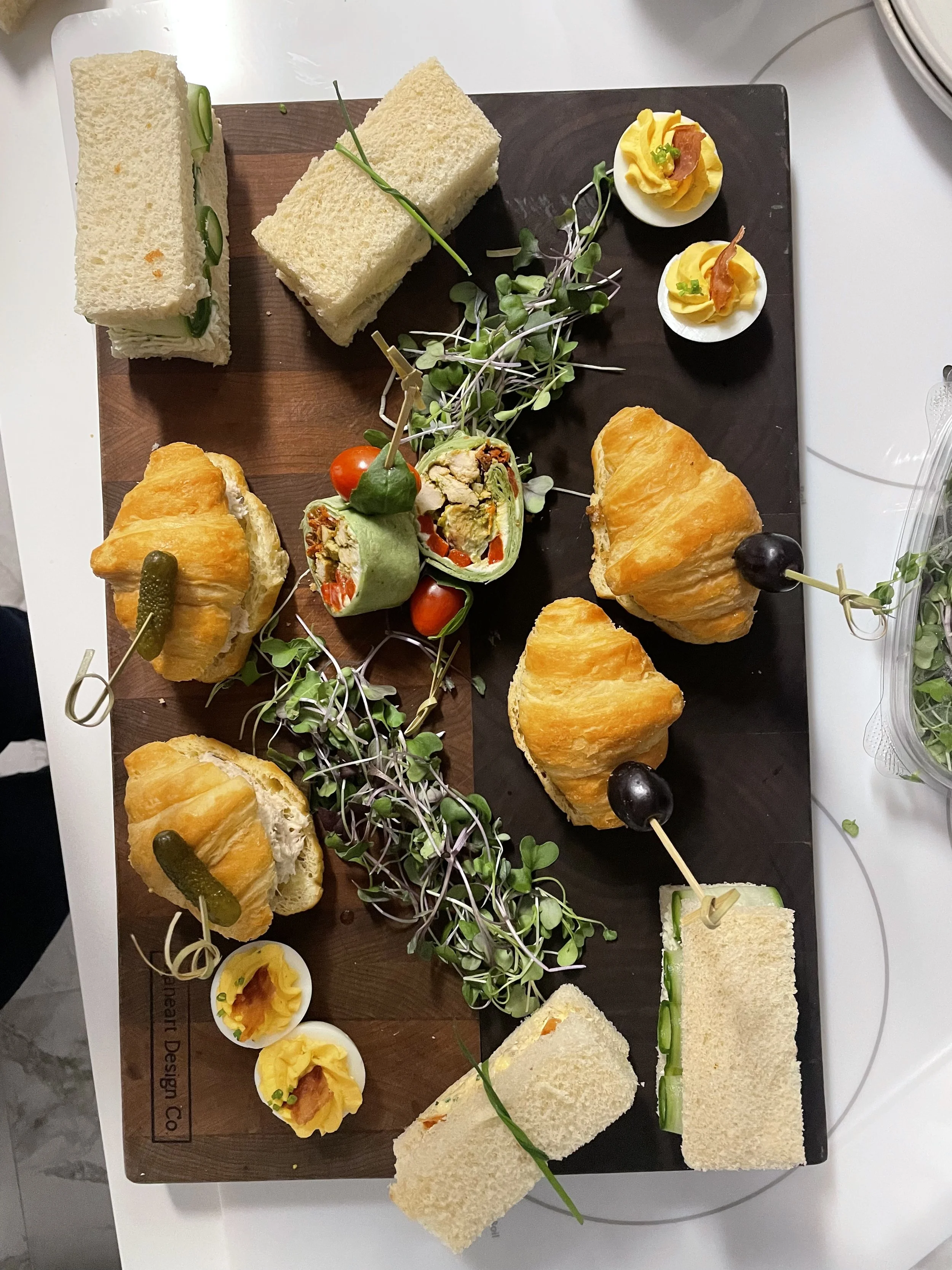 A serving tray with assorted finger foods, including sandwiches, deviled eggs, cherry tomatoes, microgreens, and skewered appetizers, arranged on a dark and wooden board.