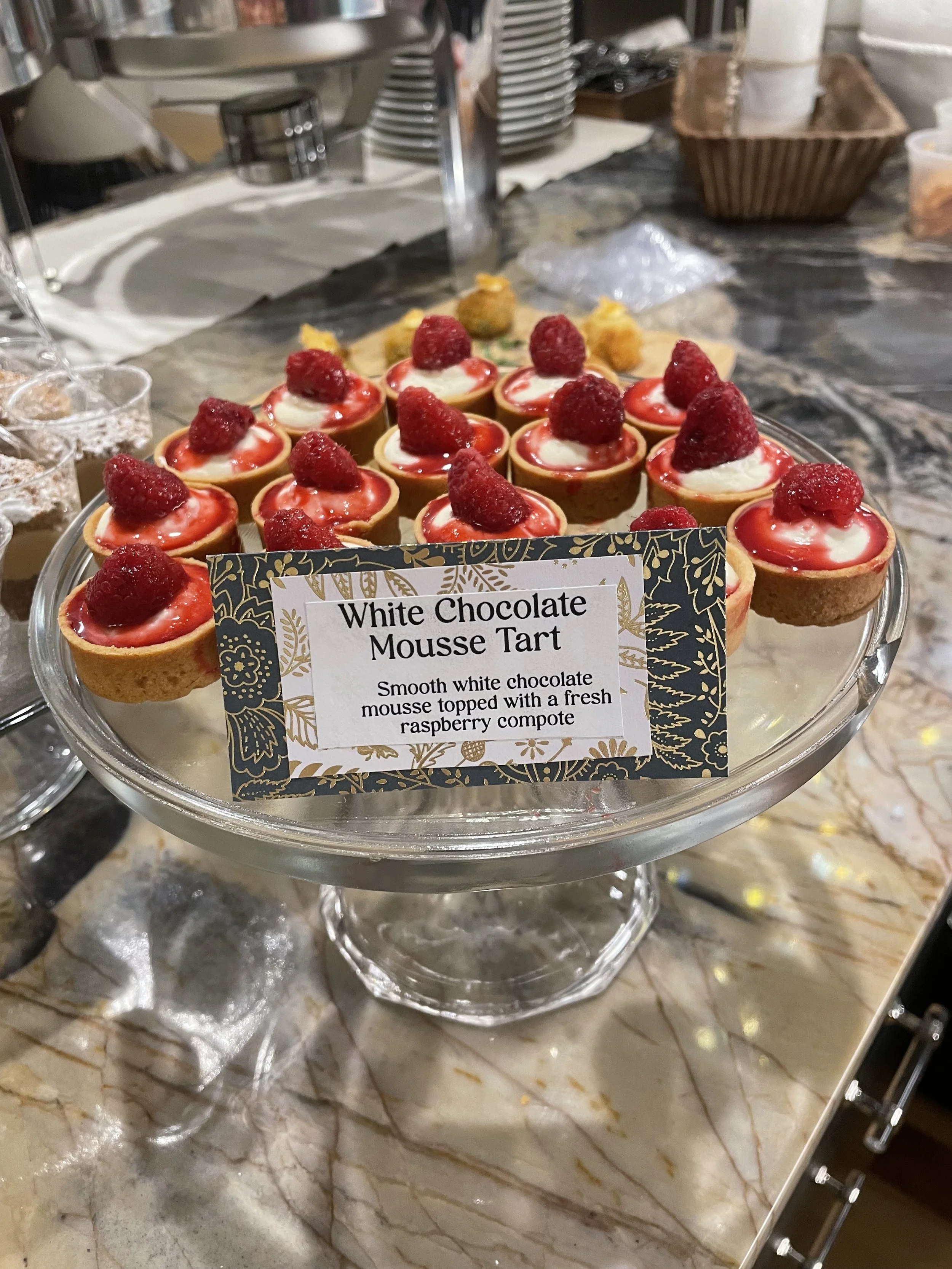 Tray of white chocolate mousse tarts topped with fresh raspberries on a glass stand at a buffet.