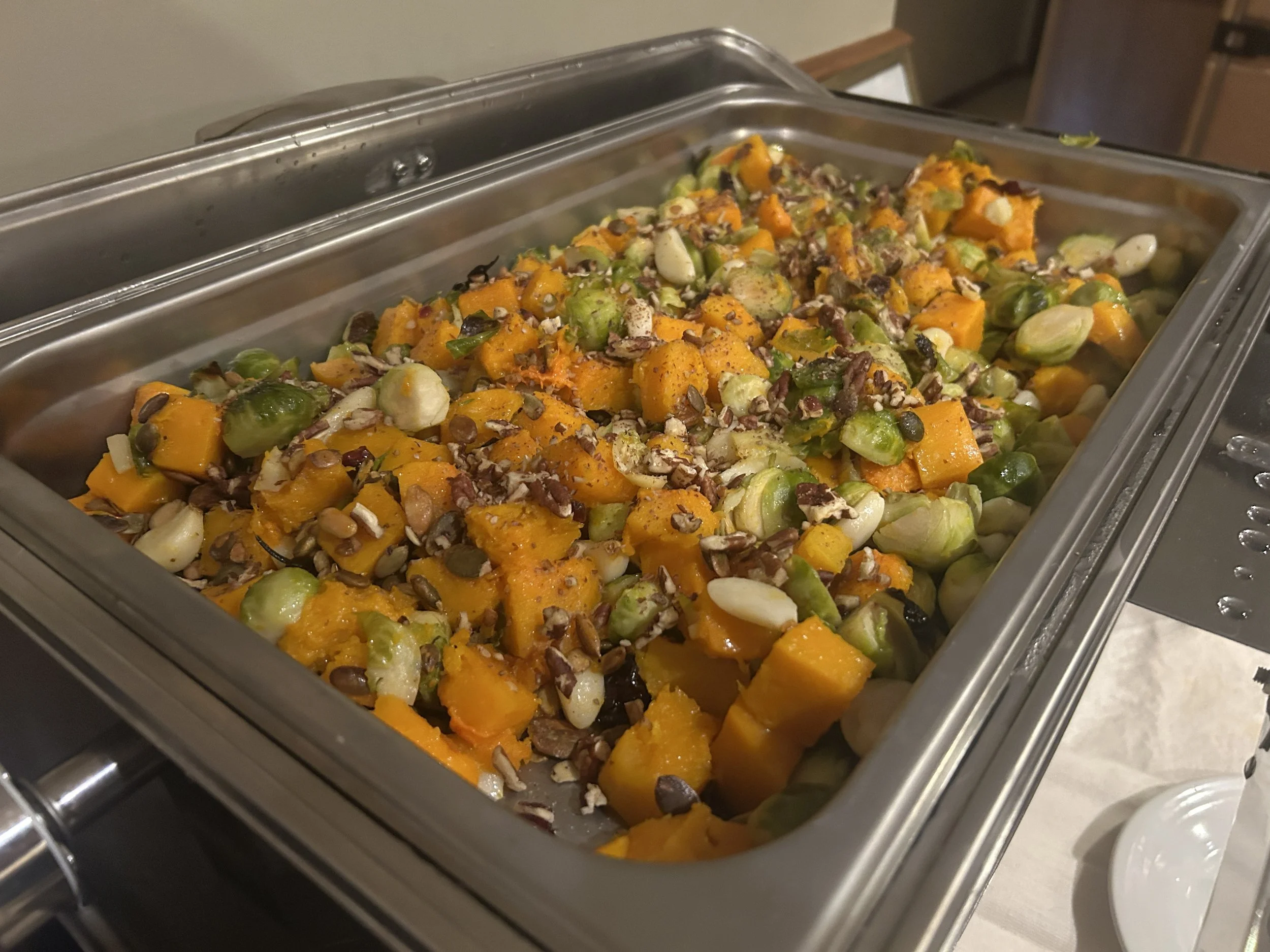 A metal chafing dish filled with roasted butternut squash, Brussels sprouts, chopped nuts, and a sprinkle of herbs.