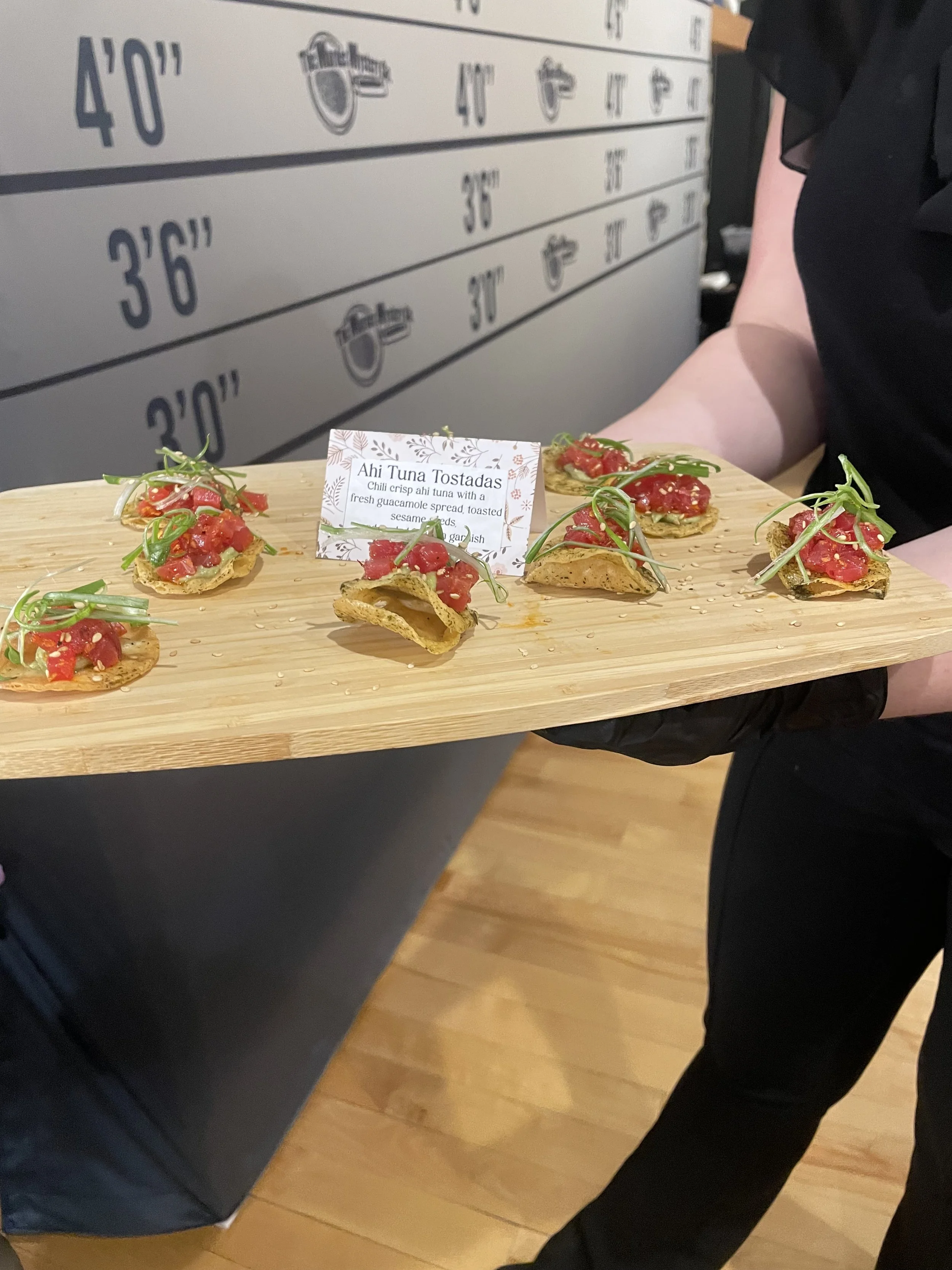 A person holding a wooden tray with small appetizer bites topped with diced tomatoes and green garnishes, with a sign that reads "Ahi Tuna Tostadas" and a description beneath.