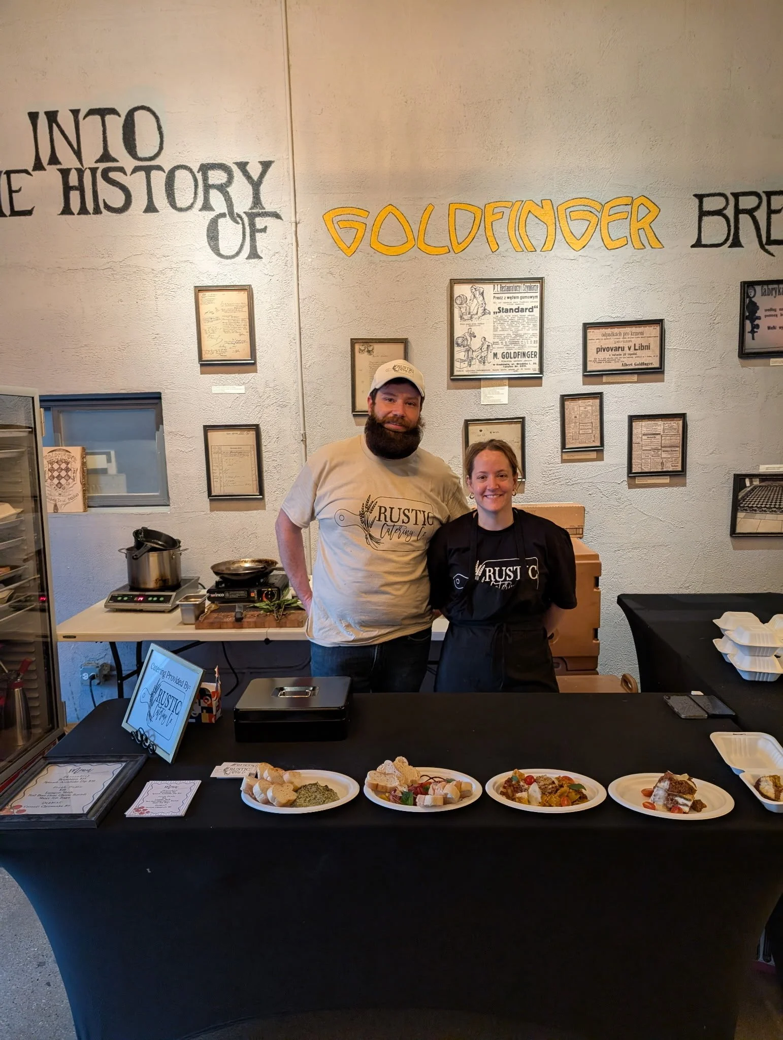 Two smiling staff members, a man and a woman, standing behind a black table with plates of food, in front of a wall with text and framed certificates. The man is wearing a beige t-shirt and a cap, the woman is wearing a black apron. The plates contain various dishes, and there is a sign indicating catering by Rustic Catering.