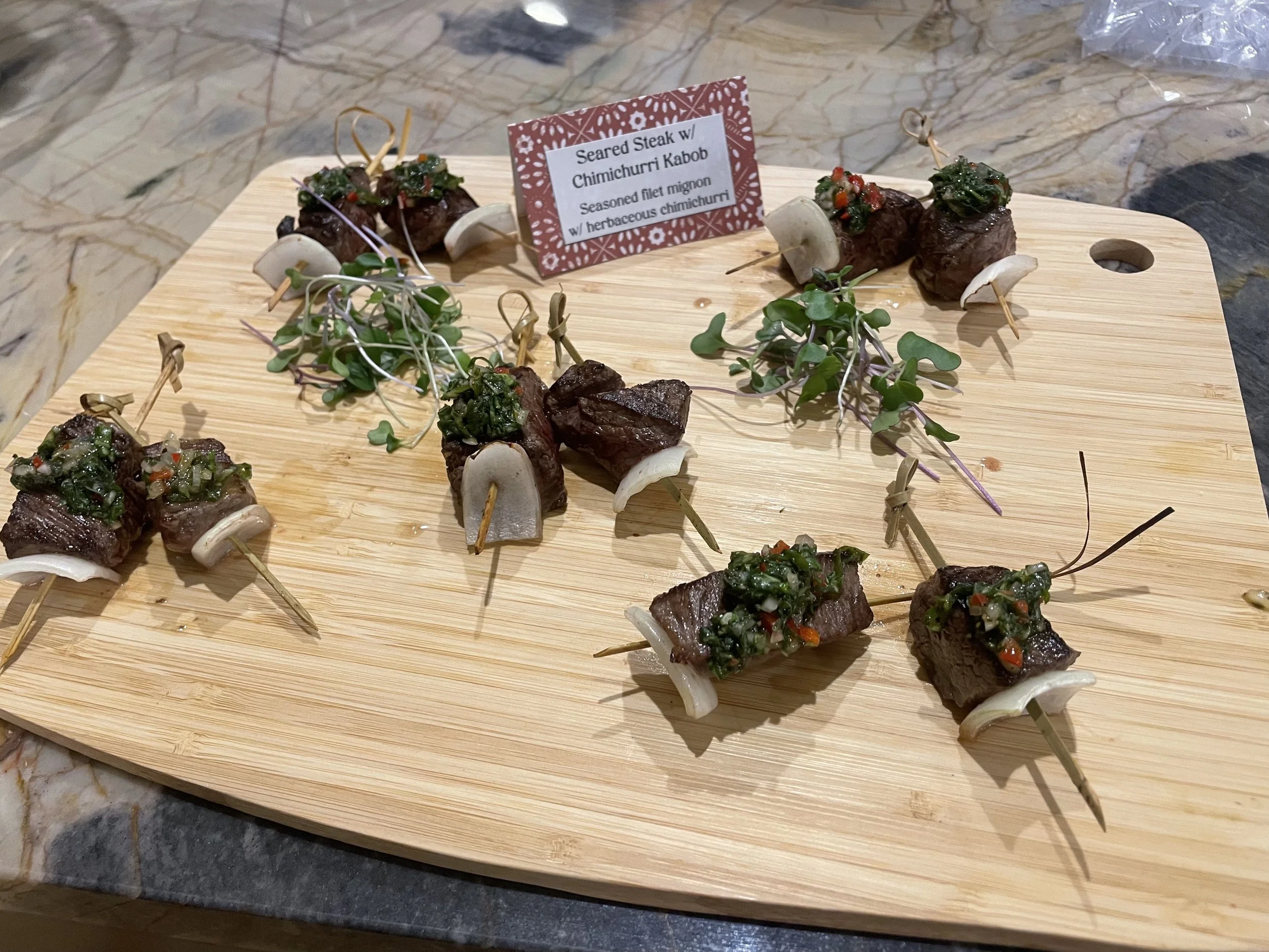 Skewers of seared steak with chimichurri sauce garnished with microgreens and sliced mushrooms on a wooden serving board.