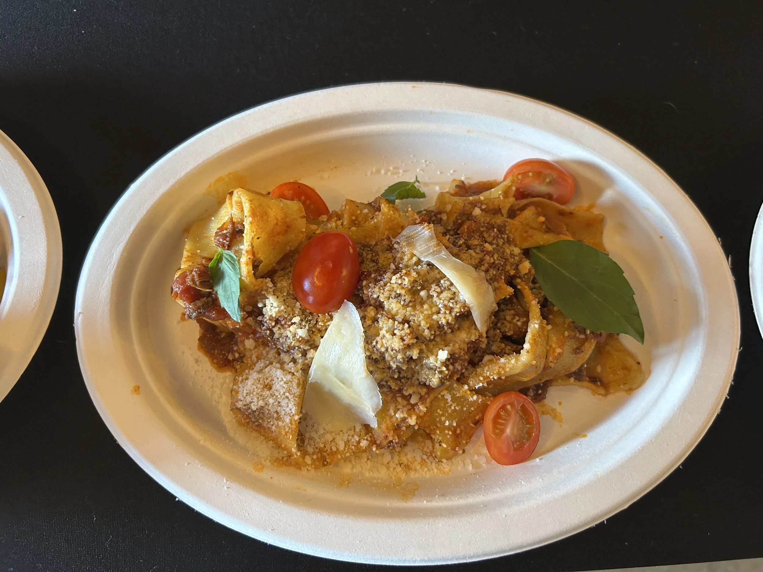 A black container with a white disposable bowl of pasta topped with cherry tomatoes, basil, grated cheese, and crushed meat sauce.