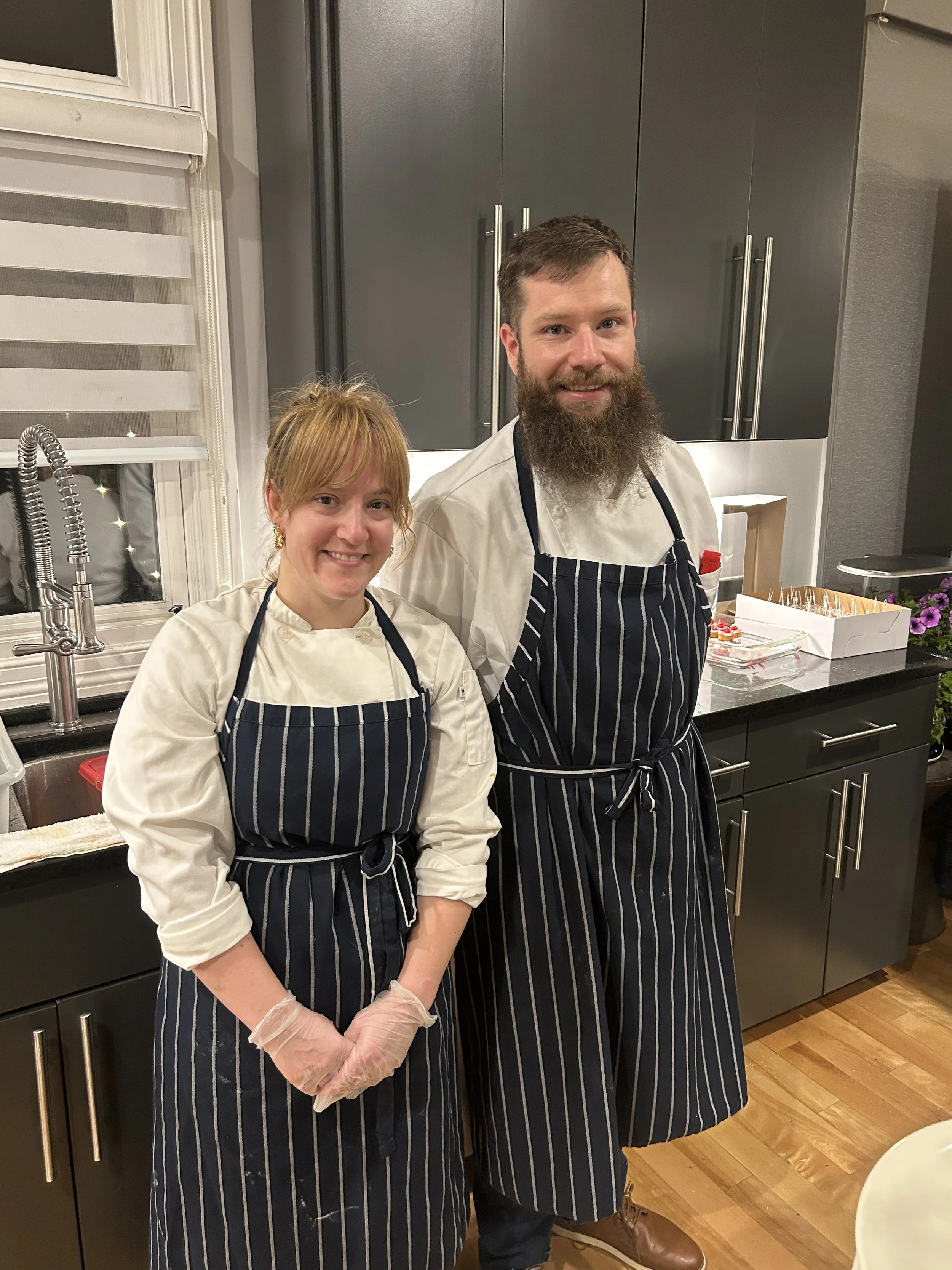 Two chefs in black and white striped aprons standing in a kitchen, smiling at the camera, with baked goods on the counter behind them.