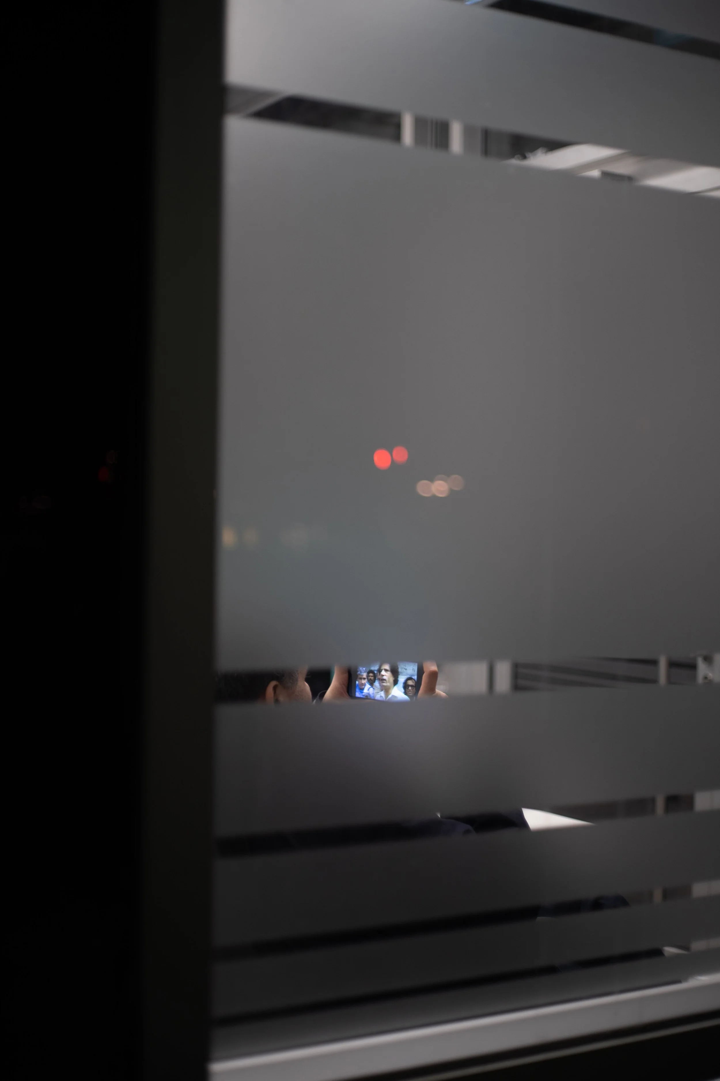 Blick durch eine bewölkte Fensterjalousie auf Menschen, die ein Smartphone fotografieren, mit roten Warnlampen im Hintergrund, bei Nacht.