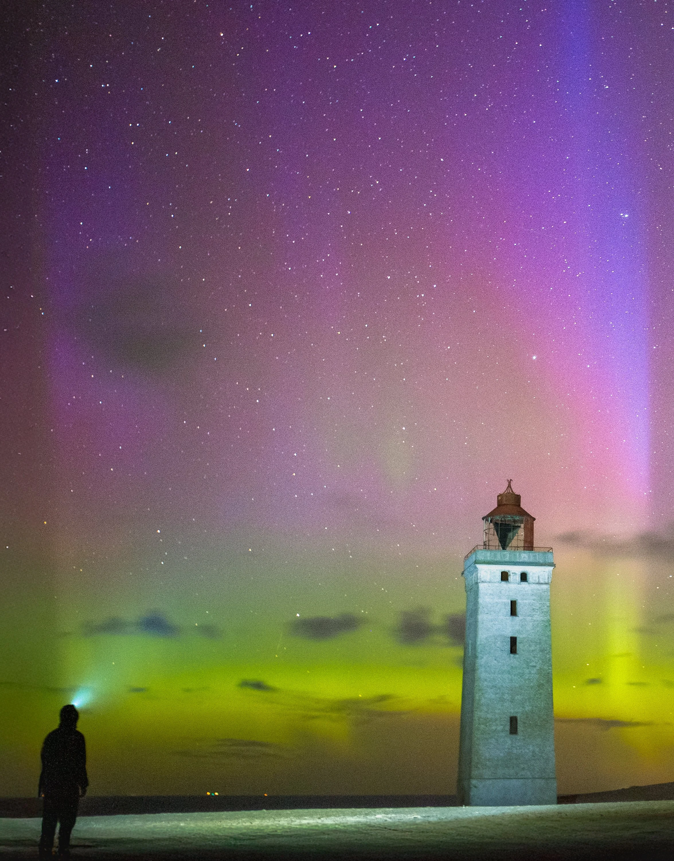 Ein Leuchtturm steht am Strand während der Nacht, der Himmel ist mit Sternen und Nordlichtern in lila, grün und gelben Farben gefüllt, mit einer Person im Vordergrund, die den Himmel betrachtet.