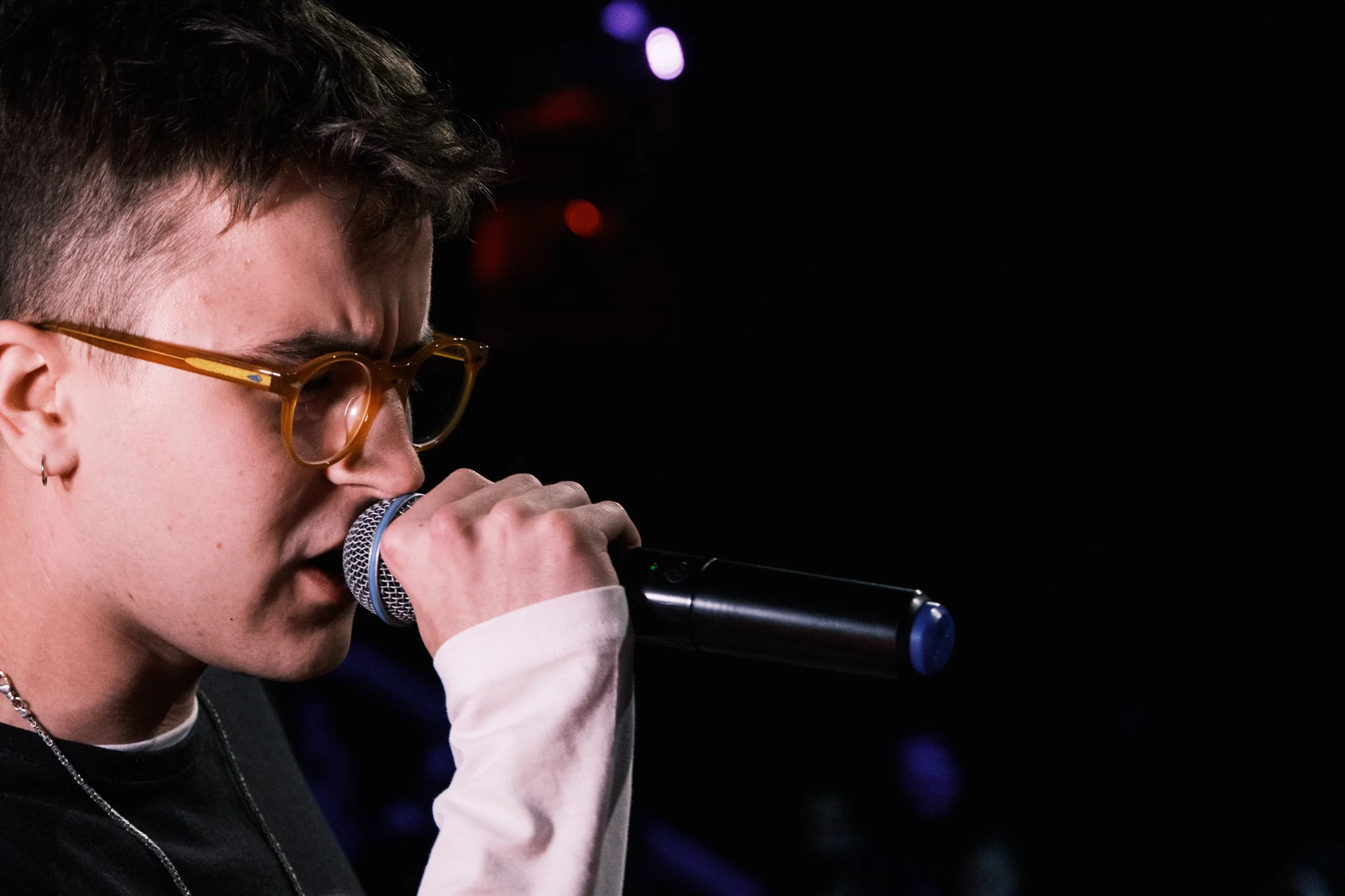 Junge Frau mit braunen Haaren, Brille und Earring singt in einem Mikrofon bei einer Veranstaltung.