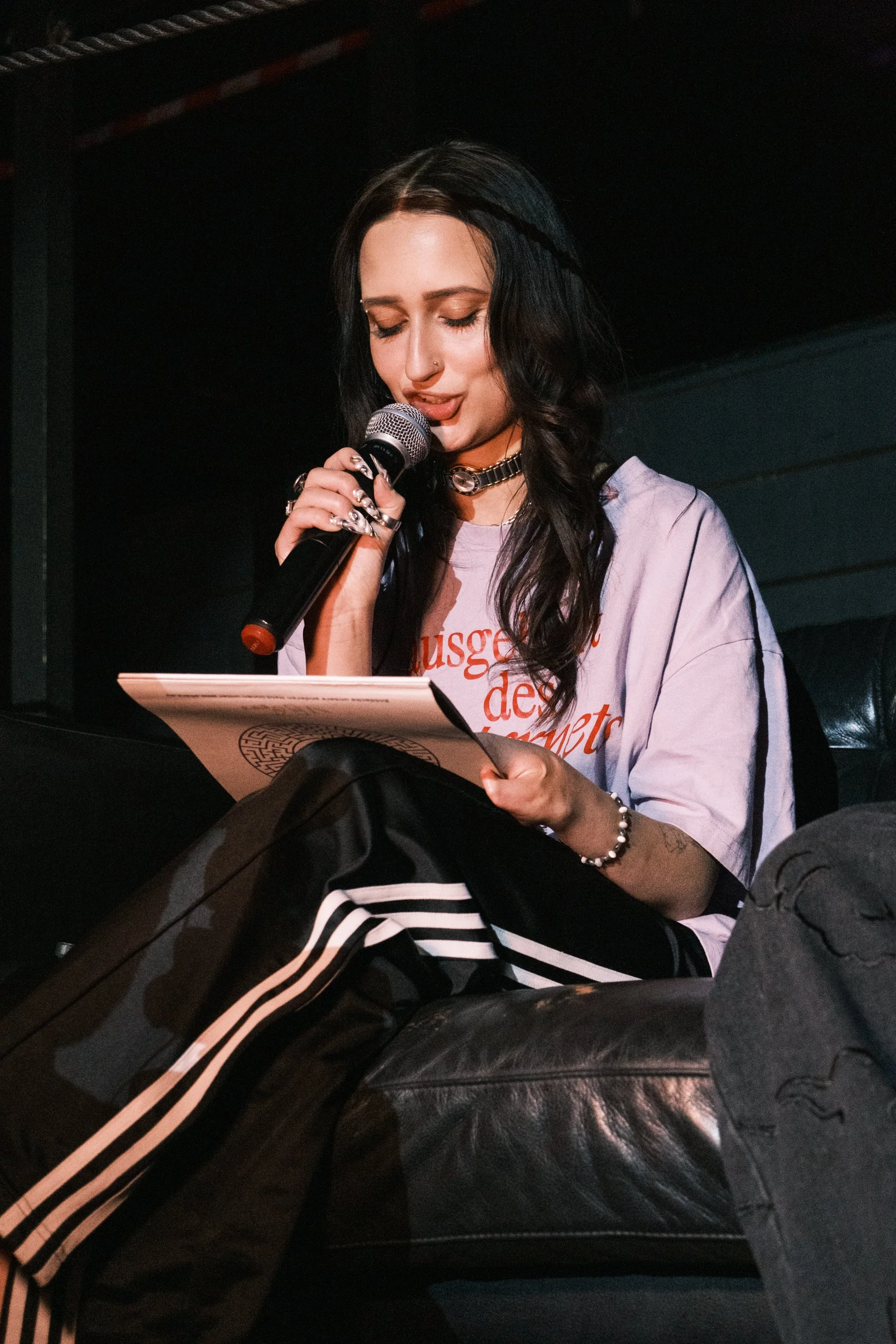 Junge Frau mit dunklen Haaren liest aus einem großen Buch mit Mikrofon in der Hand, sitzt auf einem schwarzen Sofa, trägt ein T-Shirt, Schmuck und schwarze Adidas-Sporthose mit weißen Streifen.