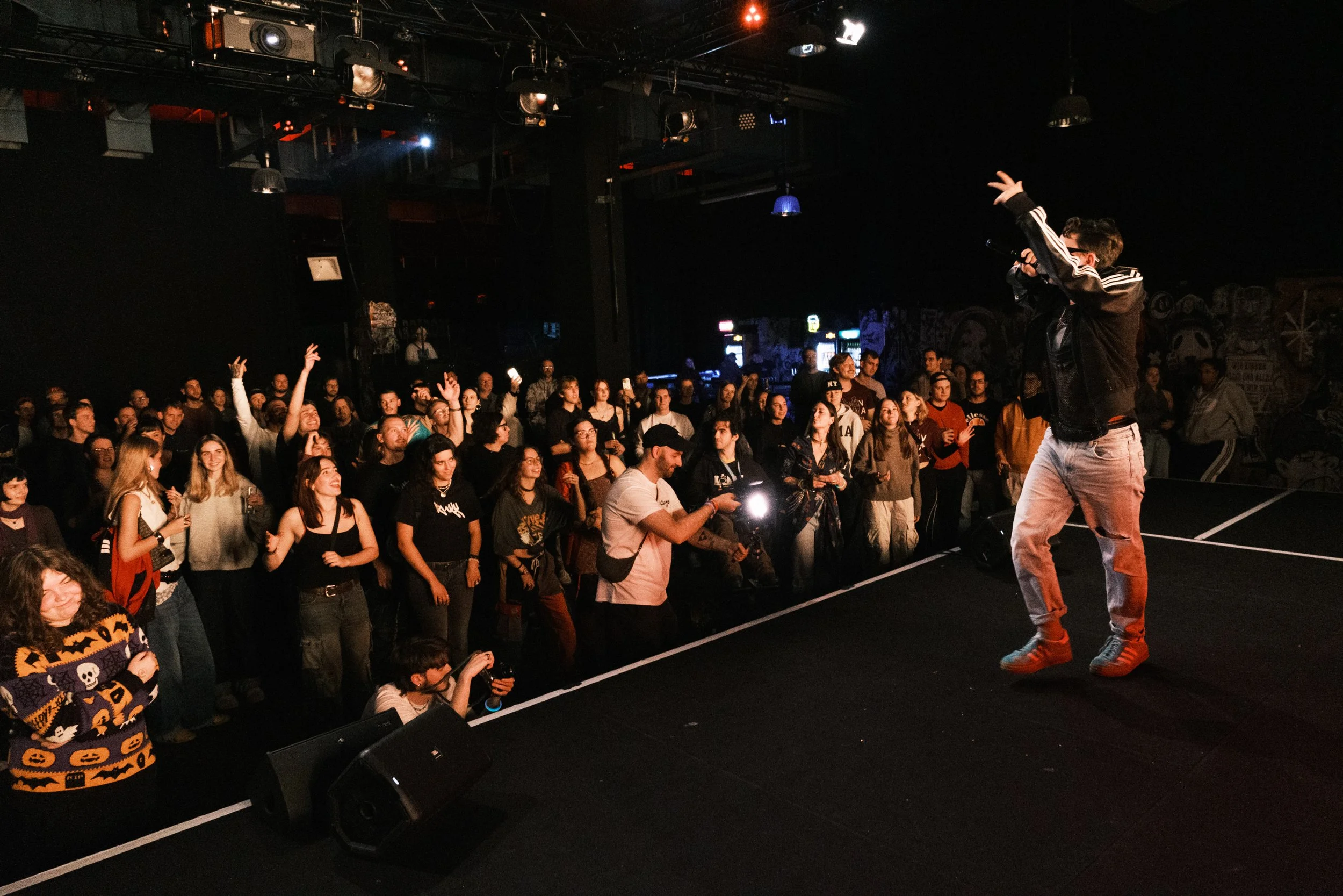 Ein Musiker auf einer Bühne während eines Konzerts, vor einem Publikum beim Singen und Tanzen im dunklen Raum mit Bühnenbeleuchtung.