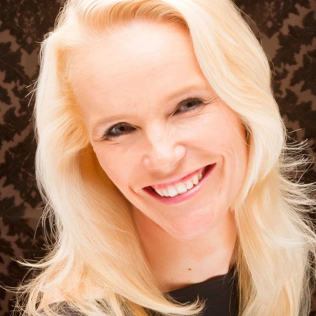 Close-up of a smiling woman with long blonde hair, light skin, and blue eyes against a dark background.