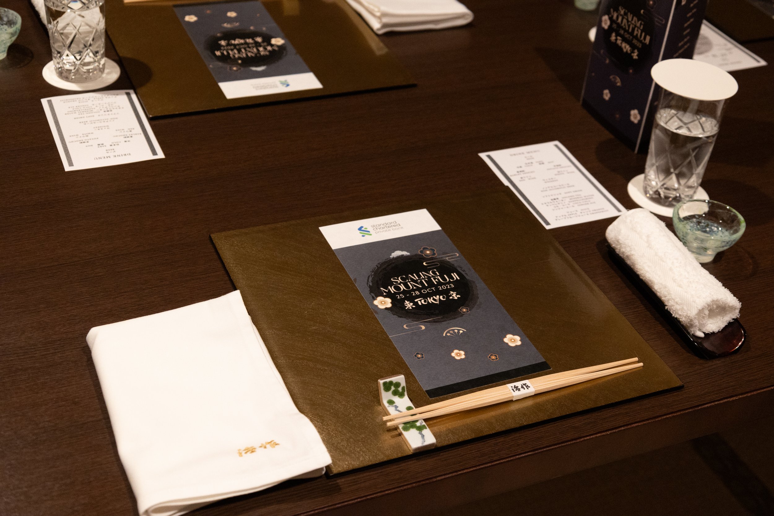 Table setting for a formal event with a menu card, chopsticks, napkin, glass of water, and a dish with a rolled towel.