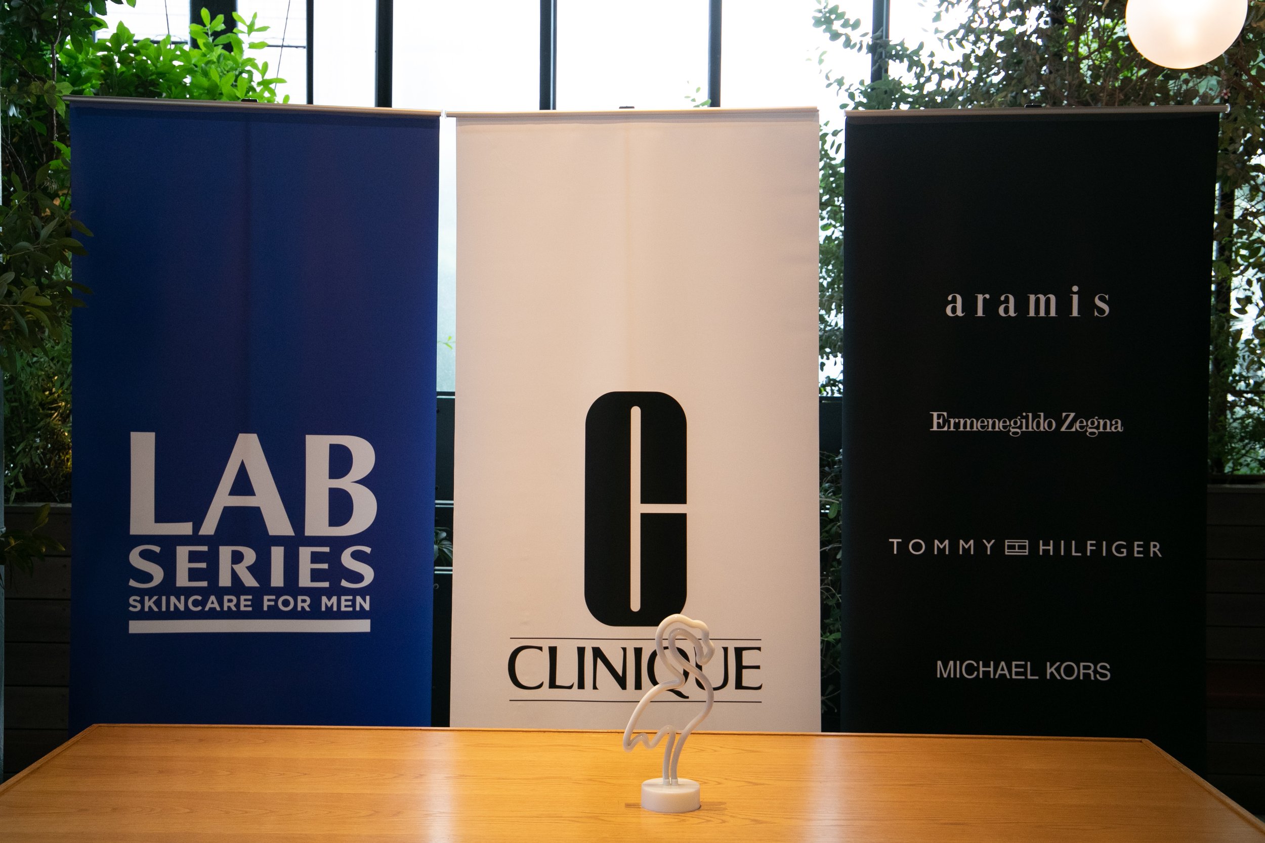 Three standing banners displaying brand logos: Lab Series Skincare for Men, Clinique, and various brands like Aramis, Ermenegildo Zegna, Tommy Hilfiger, and Michael Kors, with a small flamingo sculpture on a wooden table in front.