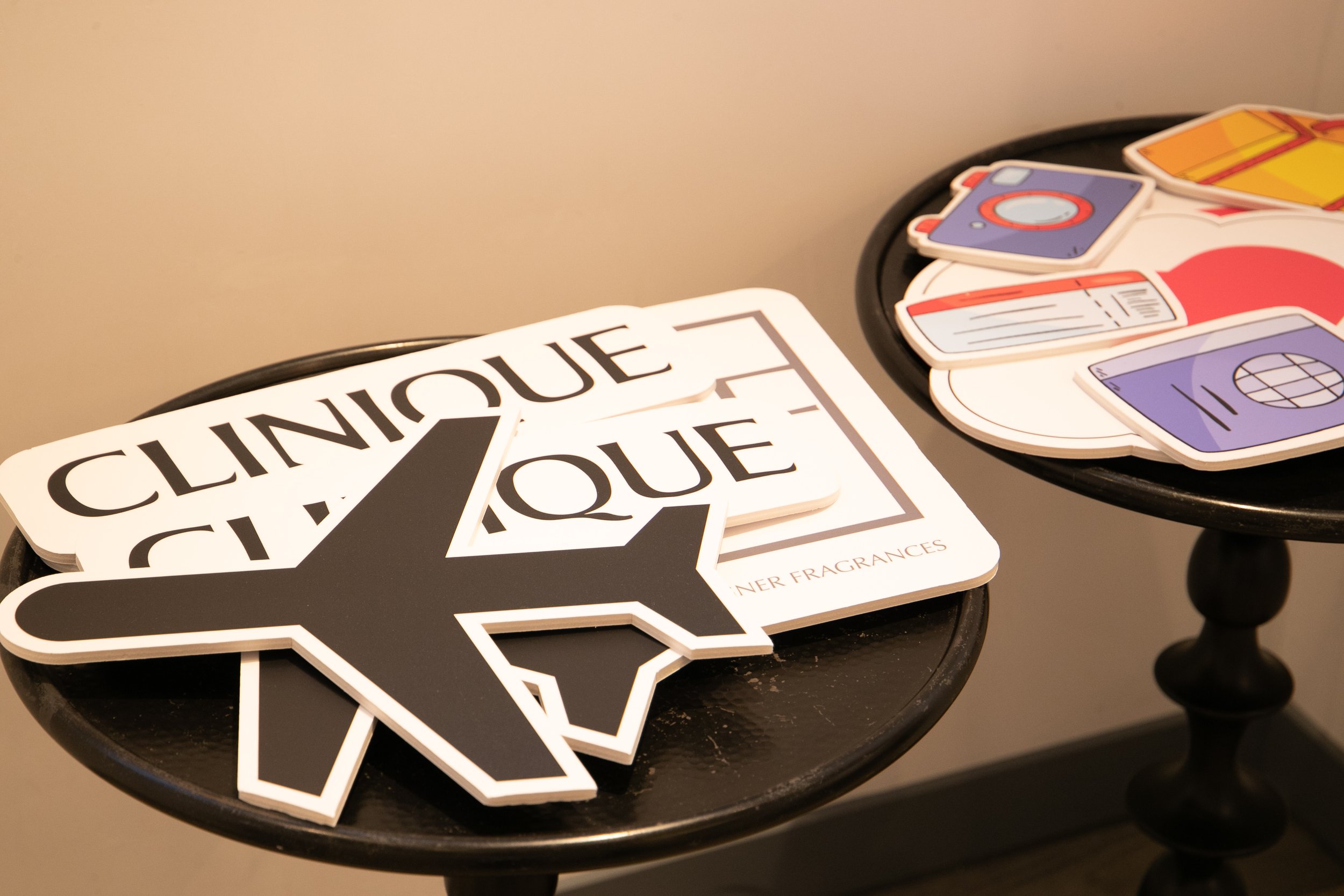 Two tables with cutout signs, including a black airplane silhouette and "Clinique" logo, and illustrations of a gift, passport, and camera on the other table.