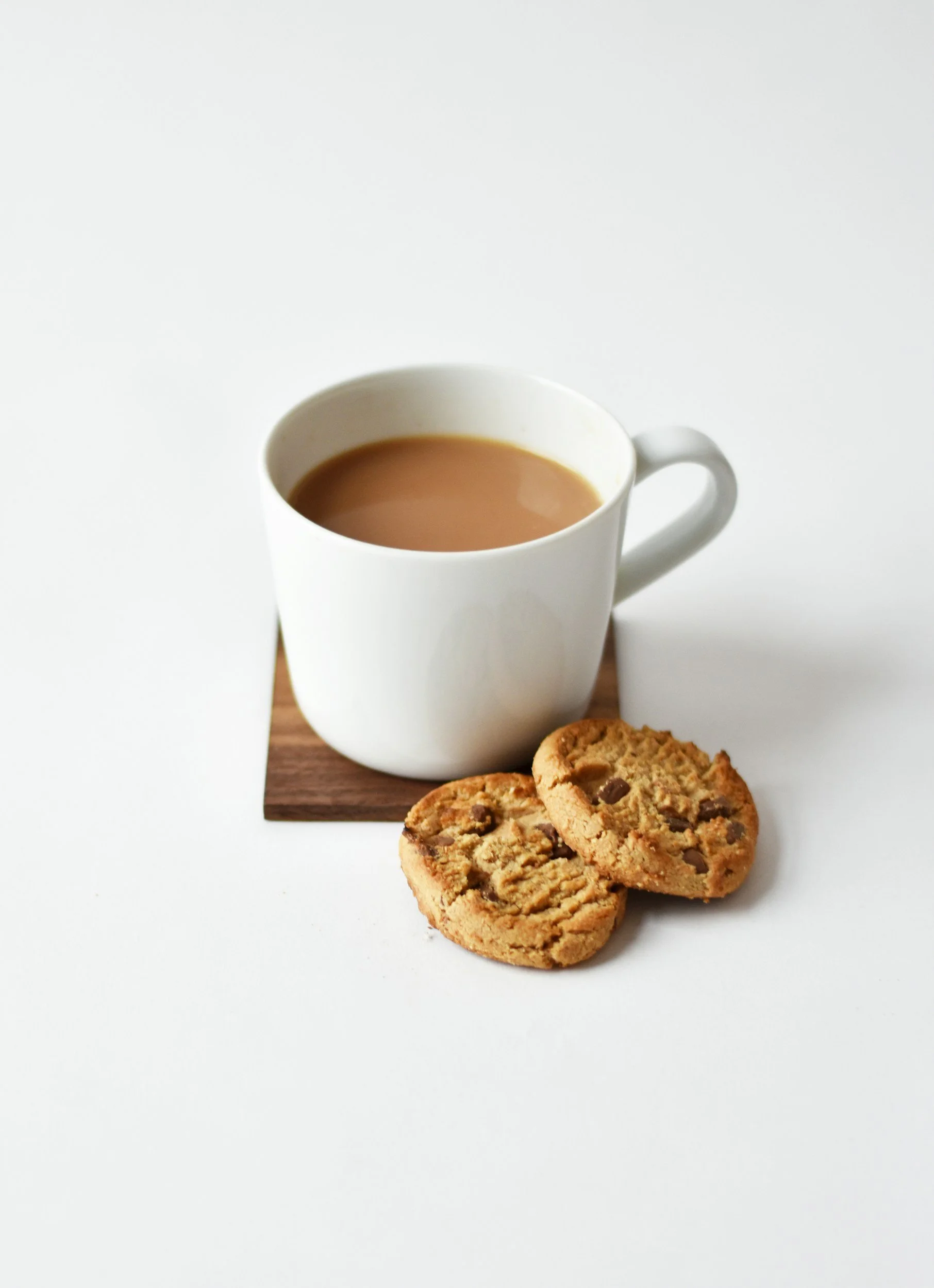 A white mug filled with coffee sitting on a wooden coaster, with two cookie biscuits beside it on a white surface.