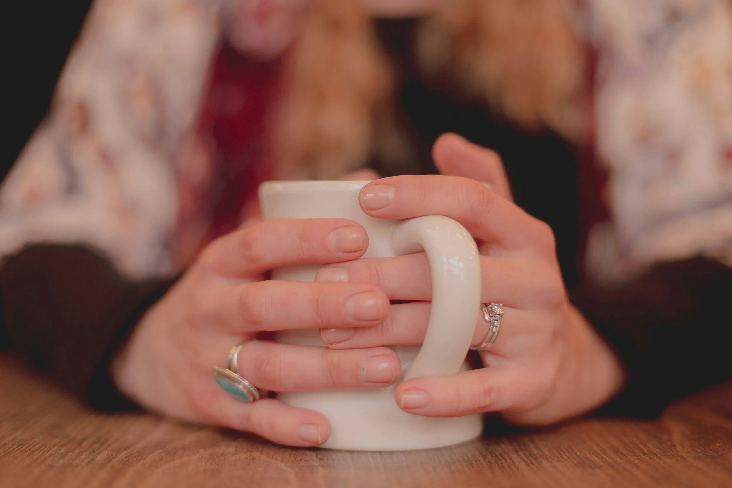Person holding a white ceramic mug with both hands, with visible rings on fingers, sitting at a wooden table.