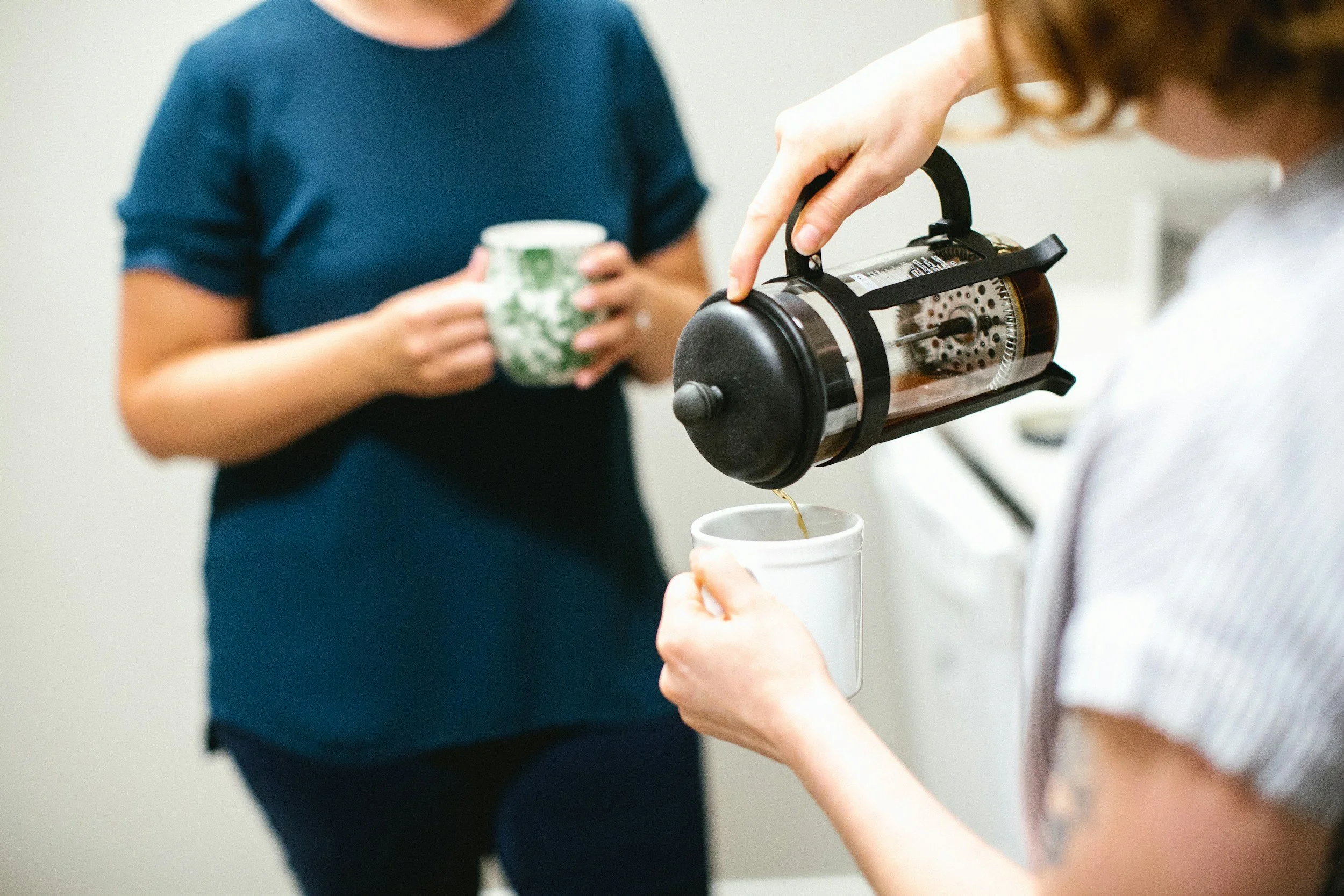 A person is pouring coffee from a French press into a white mug while another person holds a green and white patterned mug in the background.