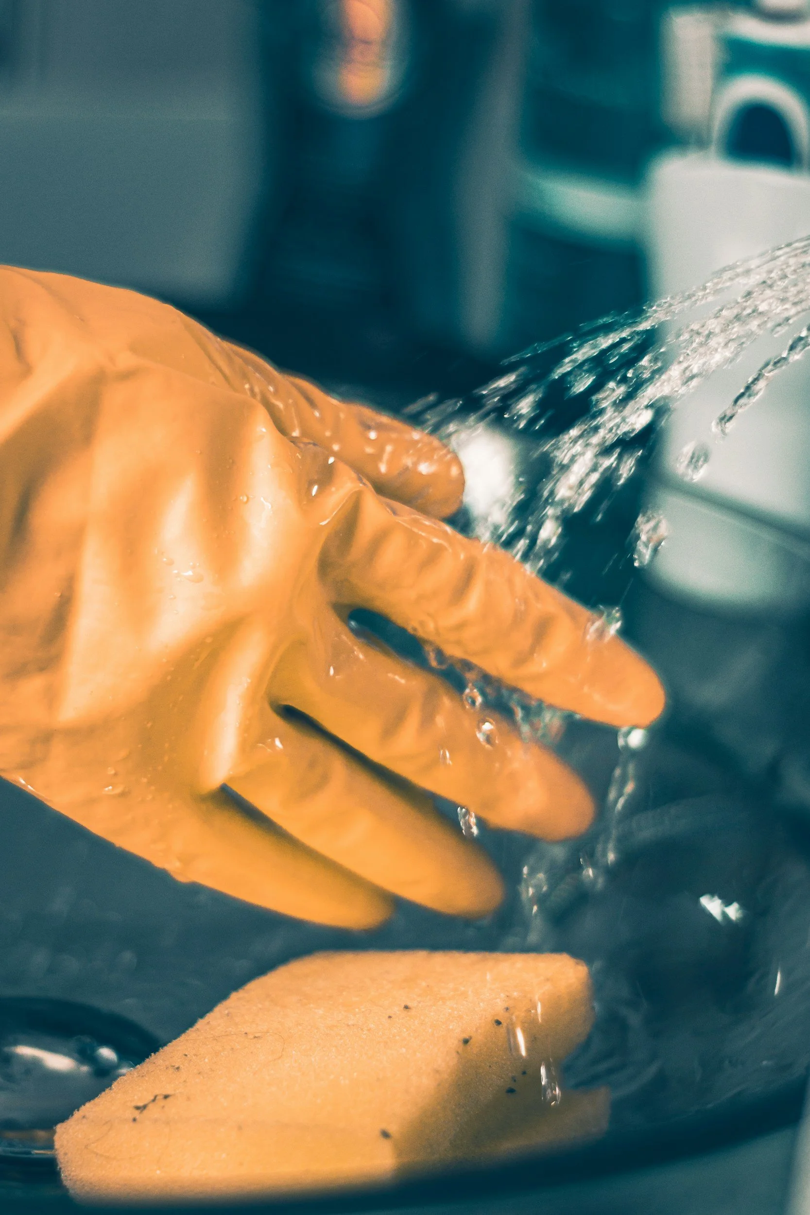 Hand wearing an orange rubber glove, rinsing a kitchen sponge under running water in a sink.
