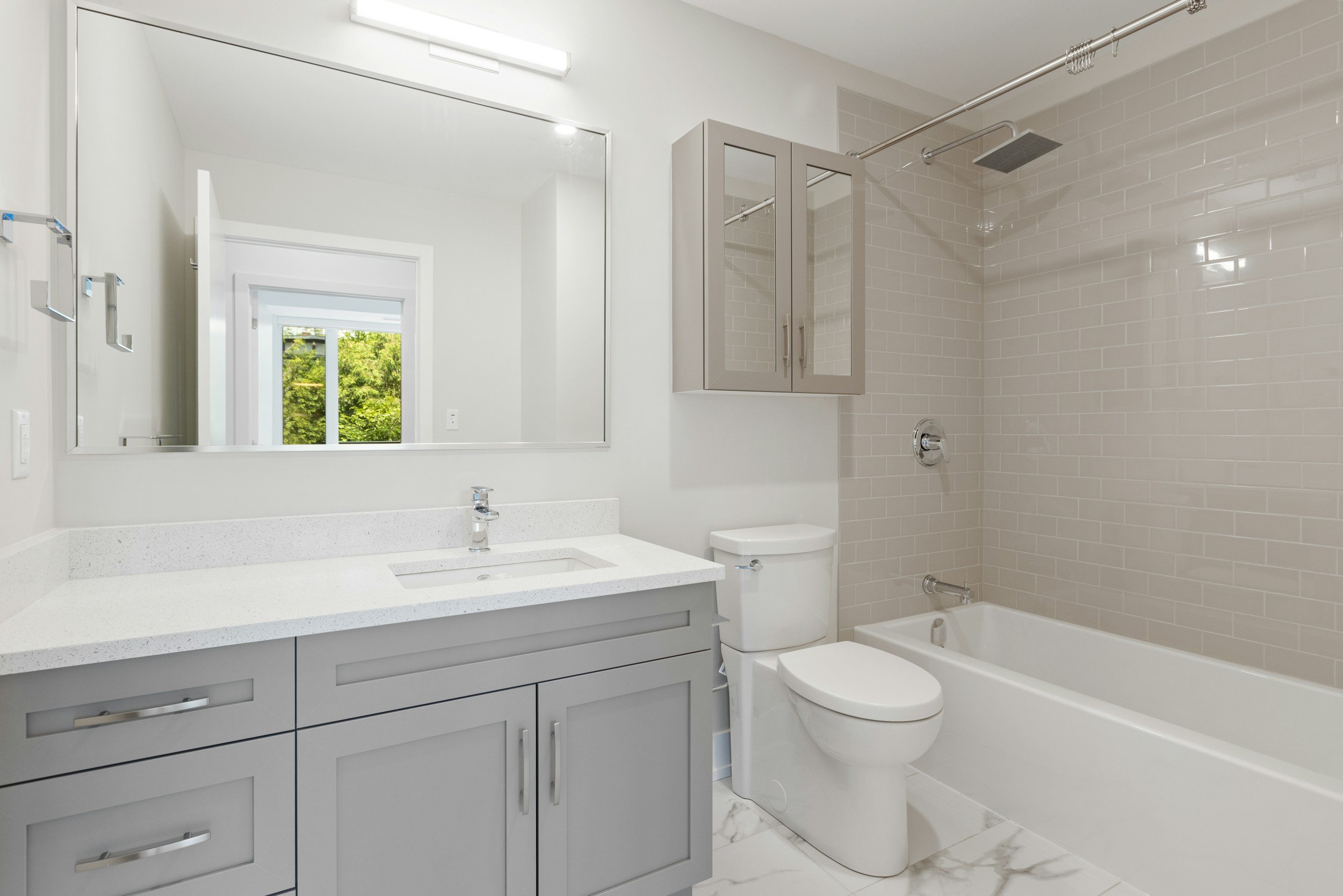Modern bathroom with a large mirror, grey vanity with white countertop, a framed mirror cabinet, a white toilet, and a bathtub with a tile wall shower.