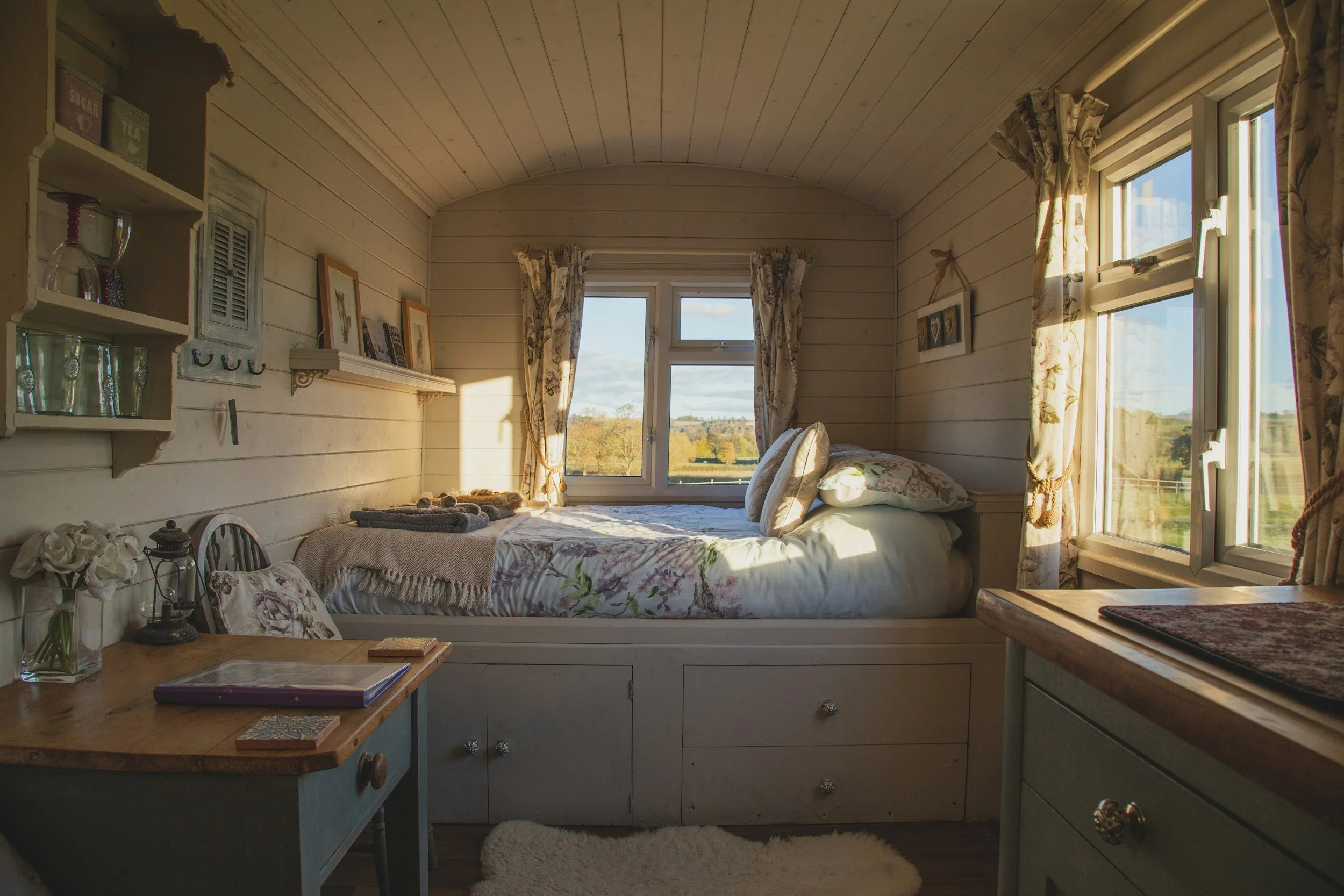Cozy bedroom with a bed, floral curtains, and natural light coming through windows, decorated with vintage and rustic style elements.