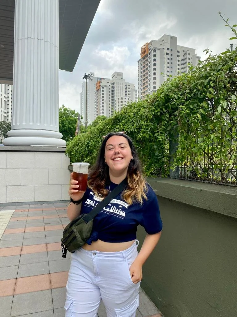A young woman smiling and holding a cup of dark soda, standing outdoors in an urban setting with high-rise buildings in the background. She is wearing a navy blue crop top, white cargo pants, and has a black shoulder bag.