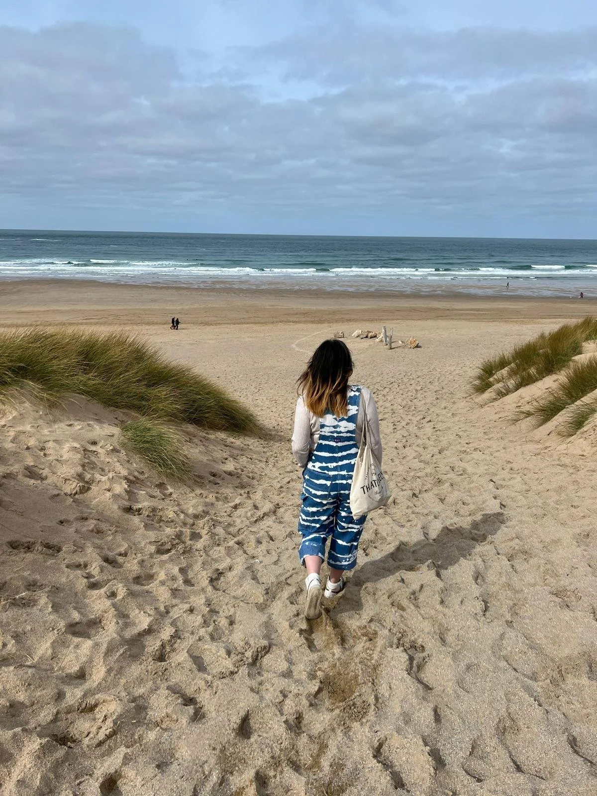 A person walking down a sandy path towards the beach, with ocean waves and cloudy sky in the background.