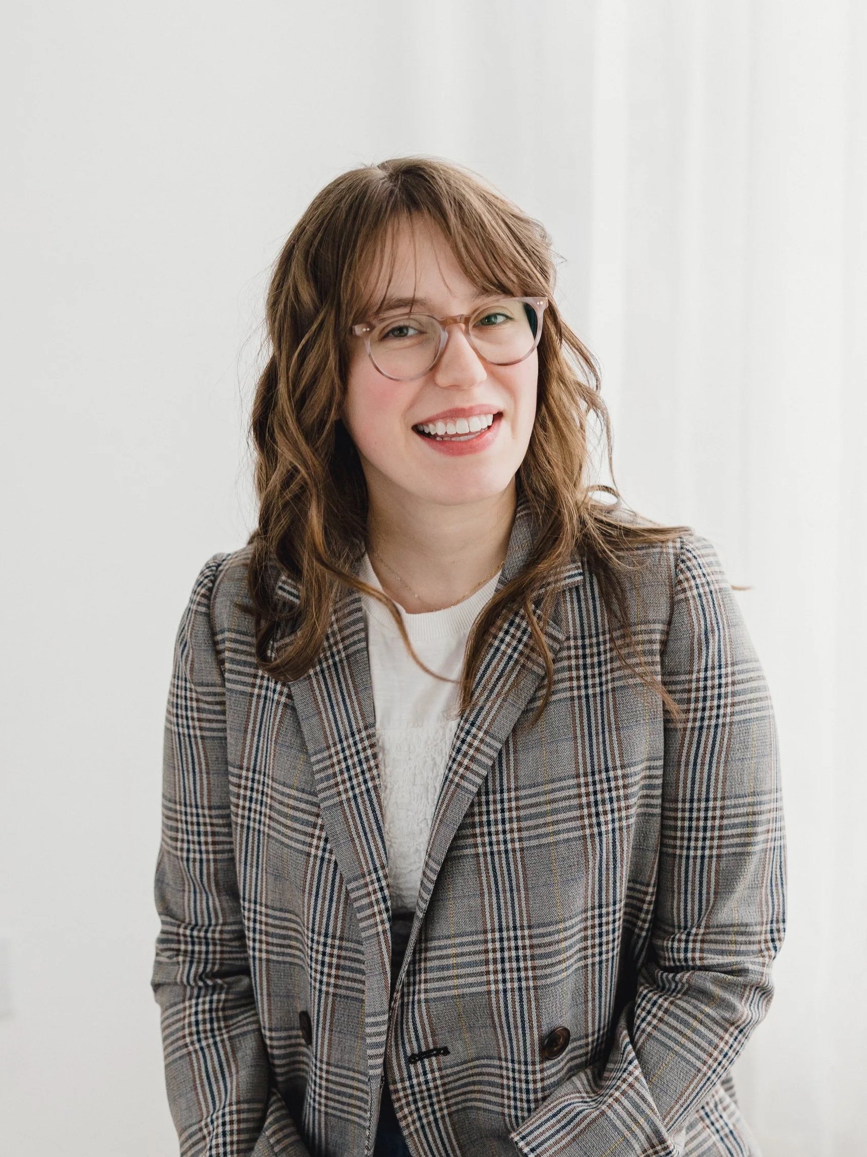 A young woman with brown wavy hair and glasses, wearing a plaid blazer, smiling.