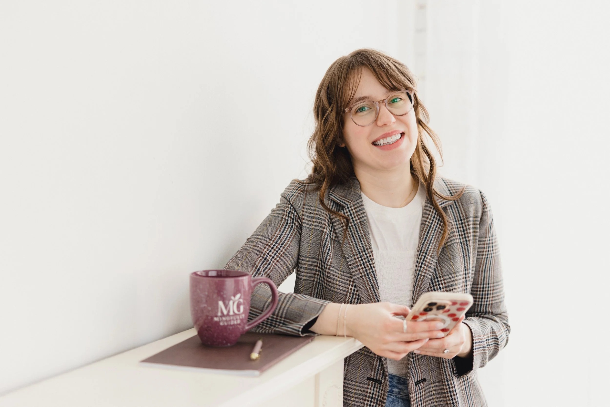 A woman with glasses, wearing a plaid blazer and white top, sitting at a white table, smiling and looking at her phone. There is a pink mug, a notebook, and a gold pen on the table.