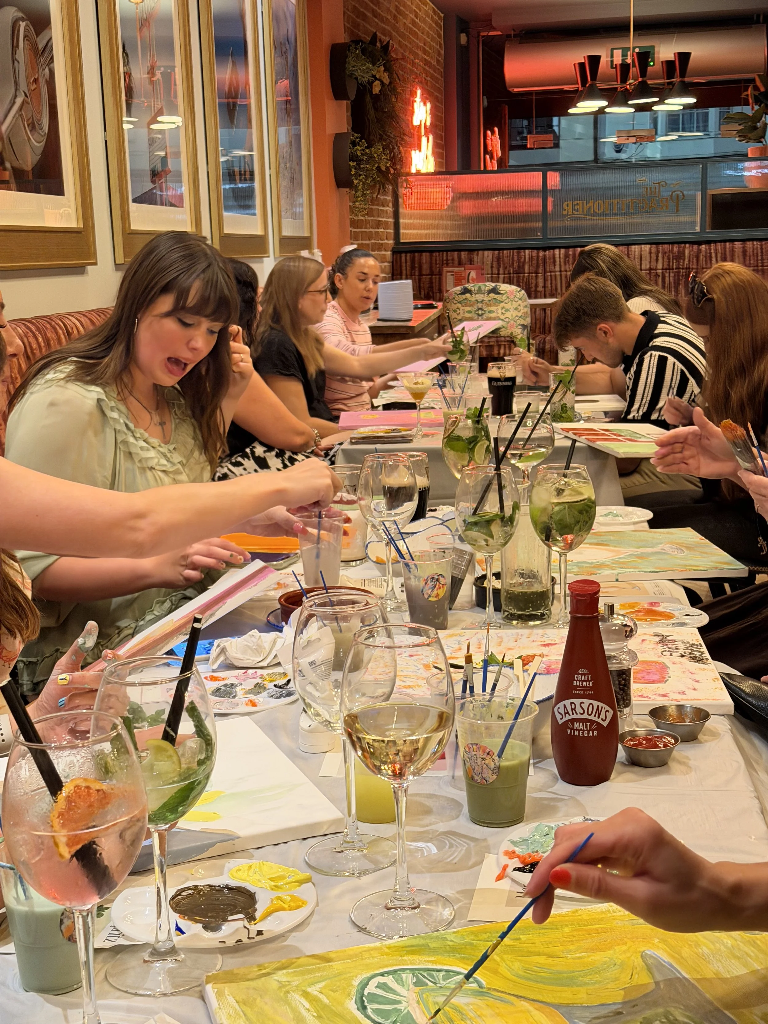 People sitting at a long table in a restaurant, painting pictures with paint, brushes, and water glasses. The background shows framed artwork, a brick wall, plants, and decorative lighting.