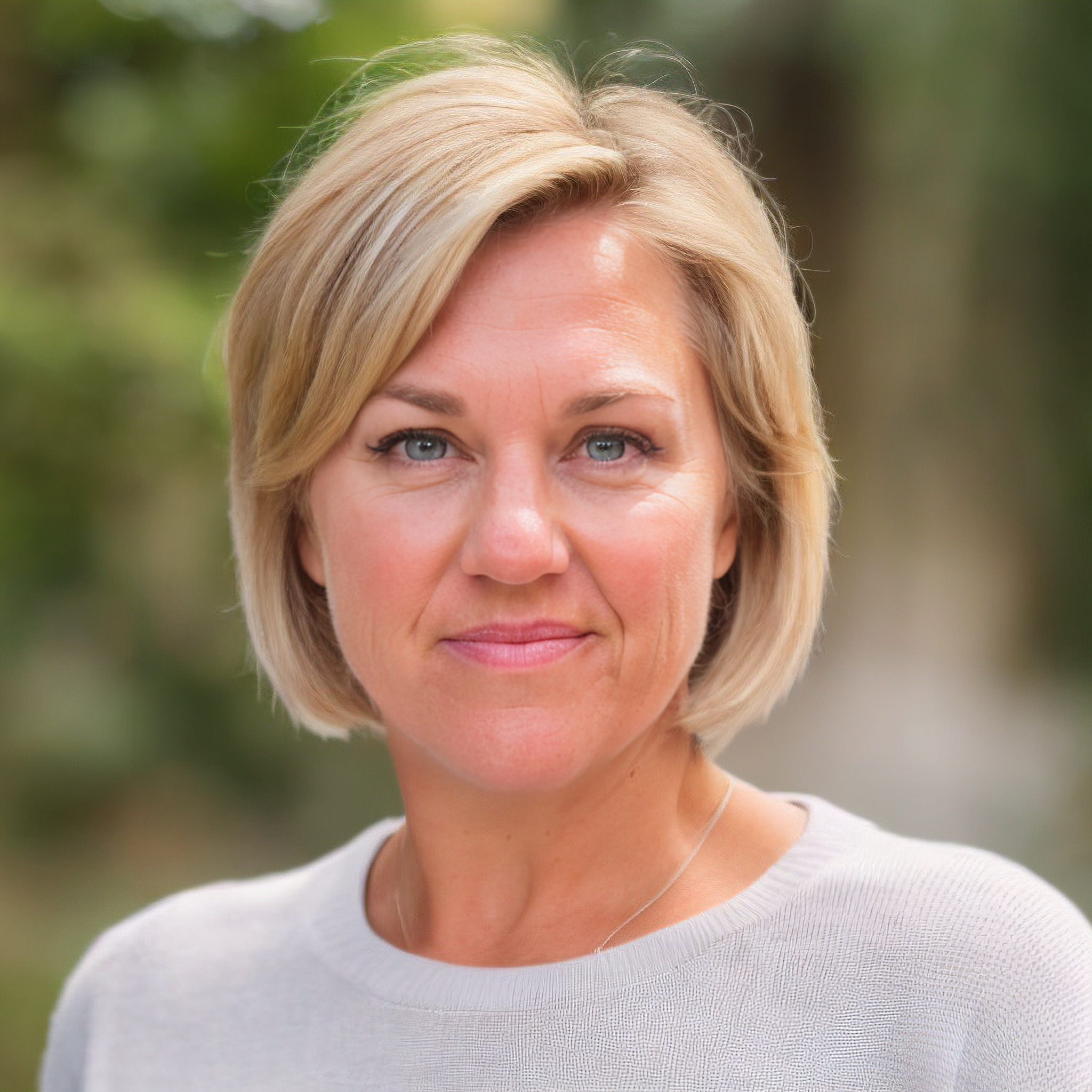 Close-up of a woman with blonde hair, blue eyes, Sarah Winslow of Locksmith Consultants, wearing a white top, in an outdoor setting with a blurred green background.