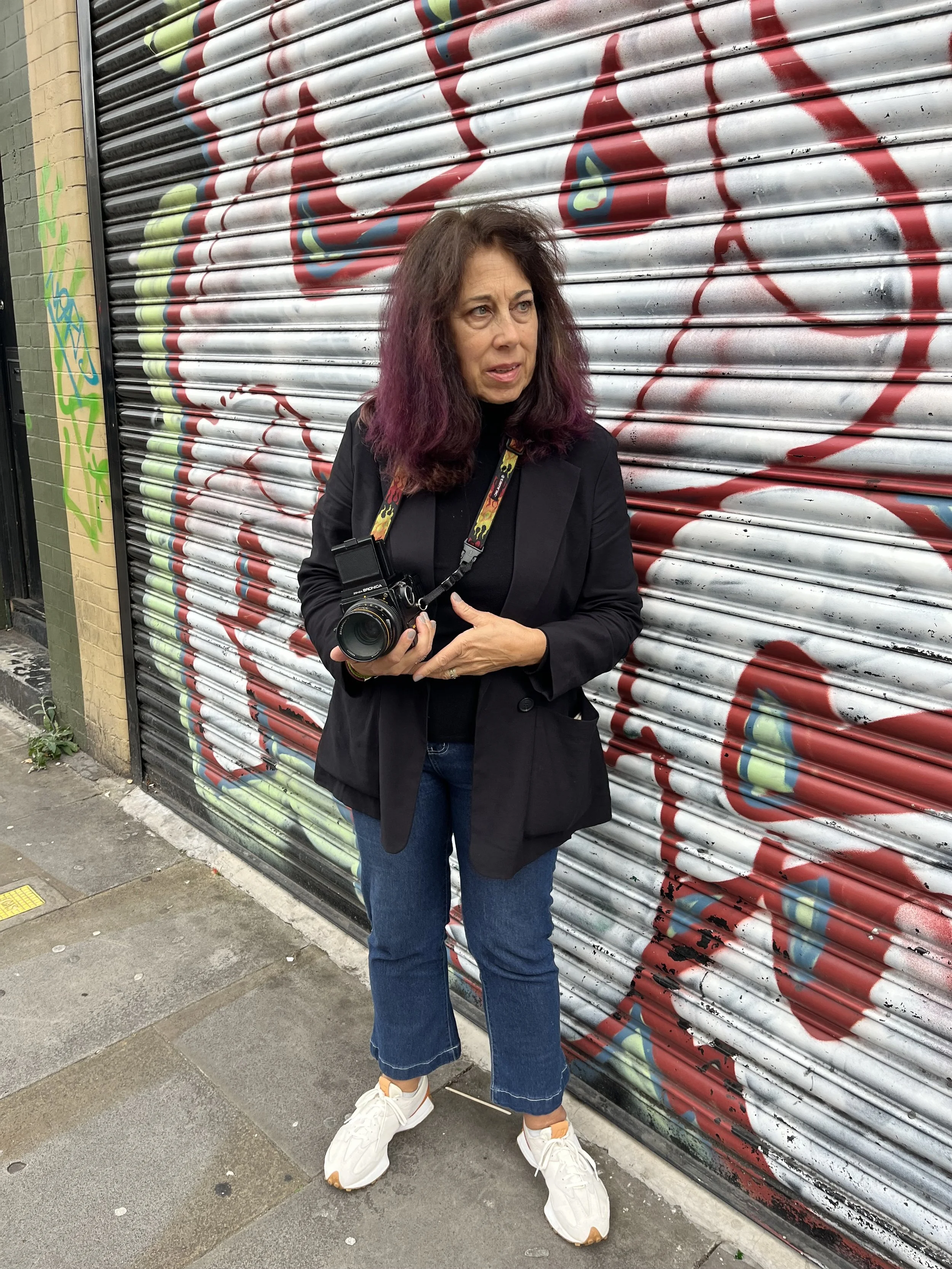 A woman with purple-tinted hair holding a camera, standing in front of a graffiti-covered metal shutter on a city sidewalk.