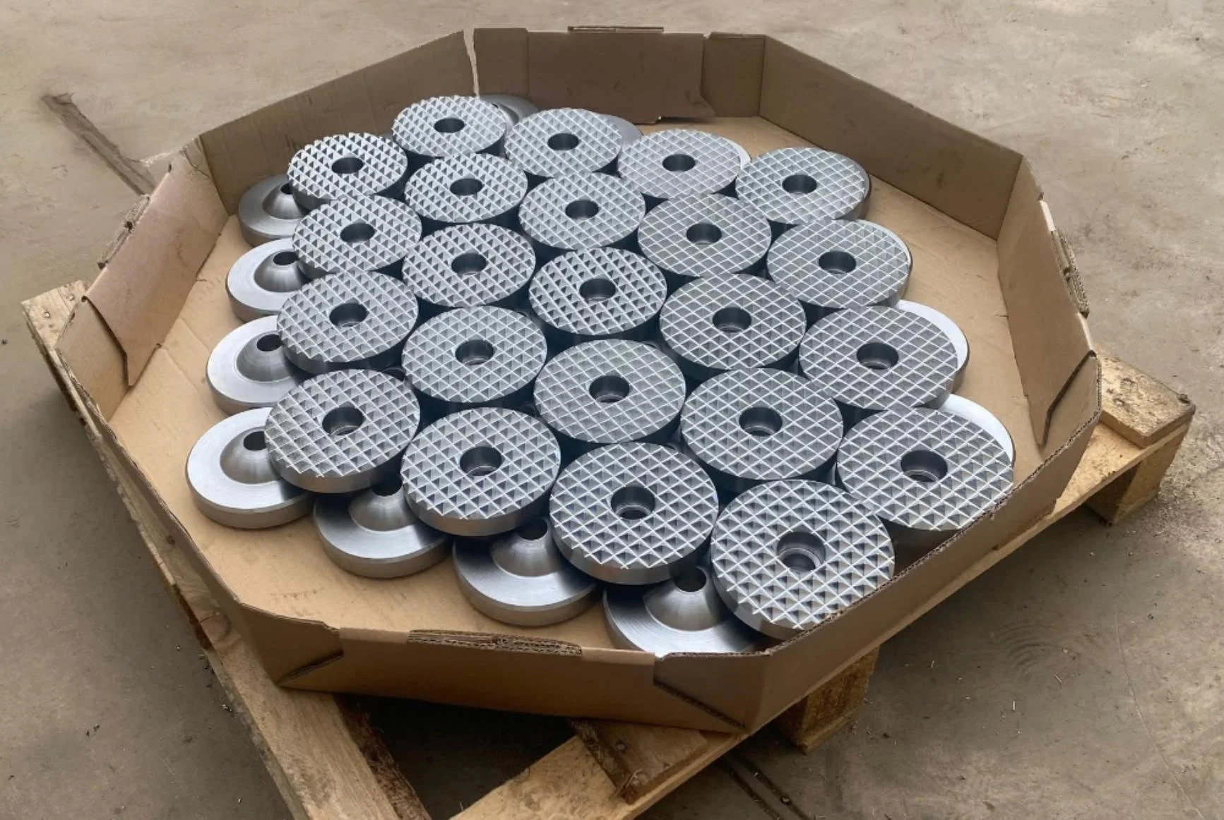 A cardboard box filled with multiple circular metal parts, some with a diamond-patterned surface and others with a smooth top, resting on a wooden pallet.