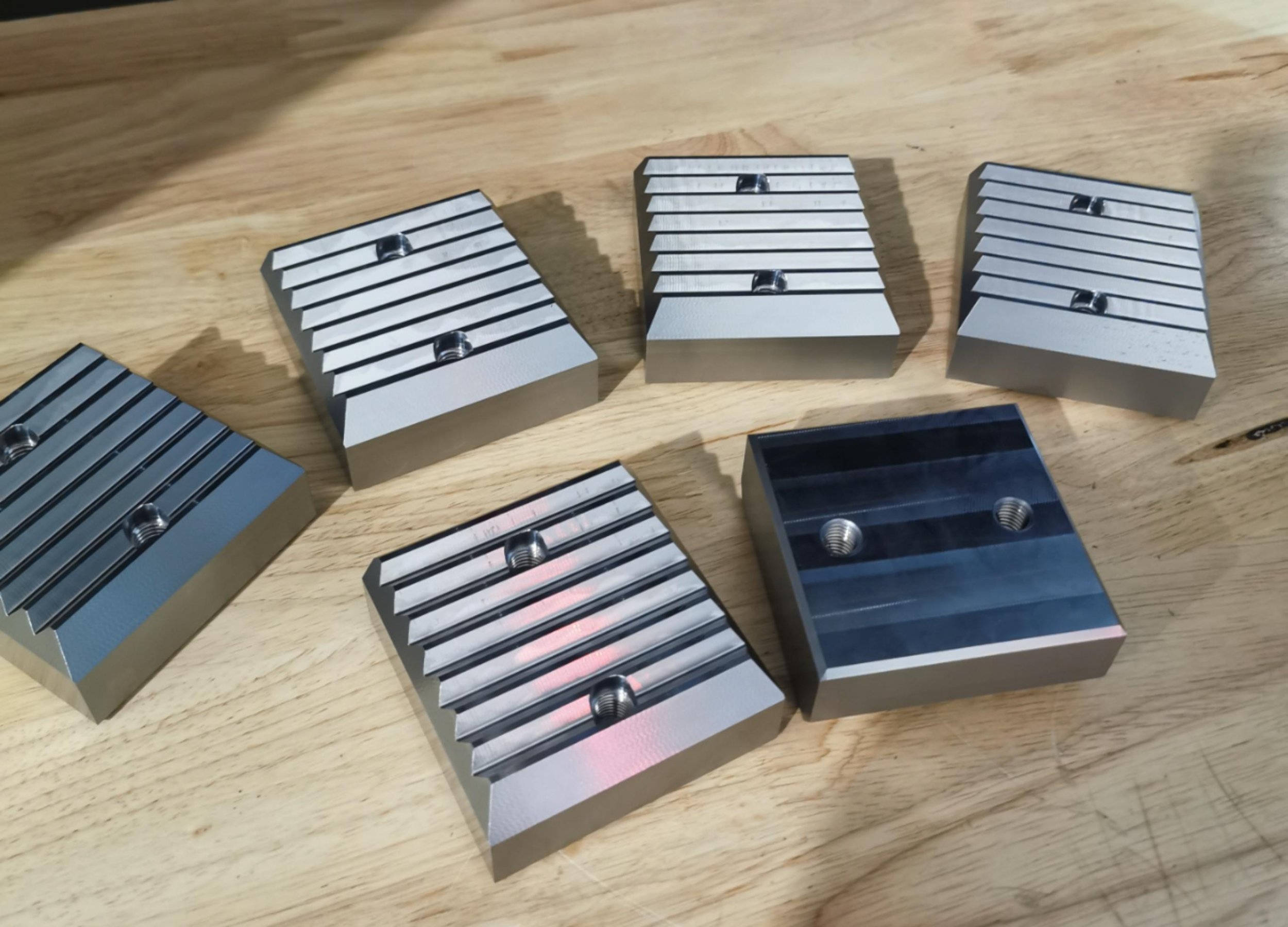 Six metallic clamps with threaded holes on a wooden surface, used for holding objects securely during machining or assembly.