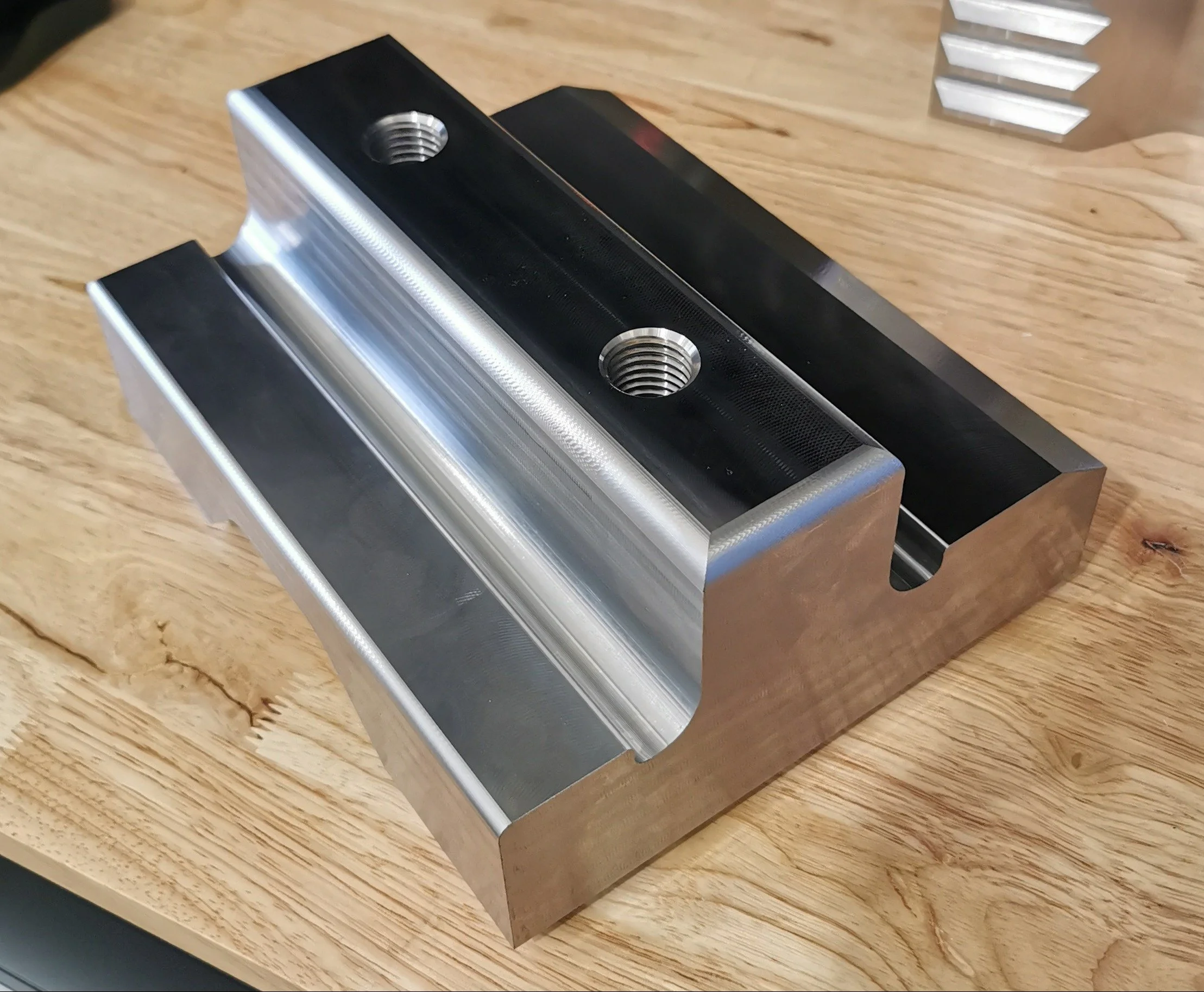 A machined metal block with threaded holes on top, placed on a wooden surface.