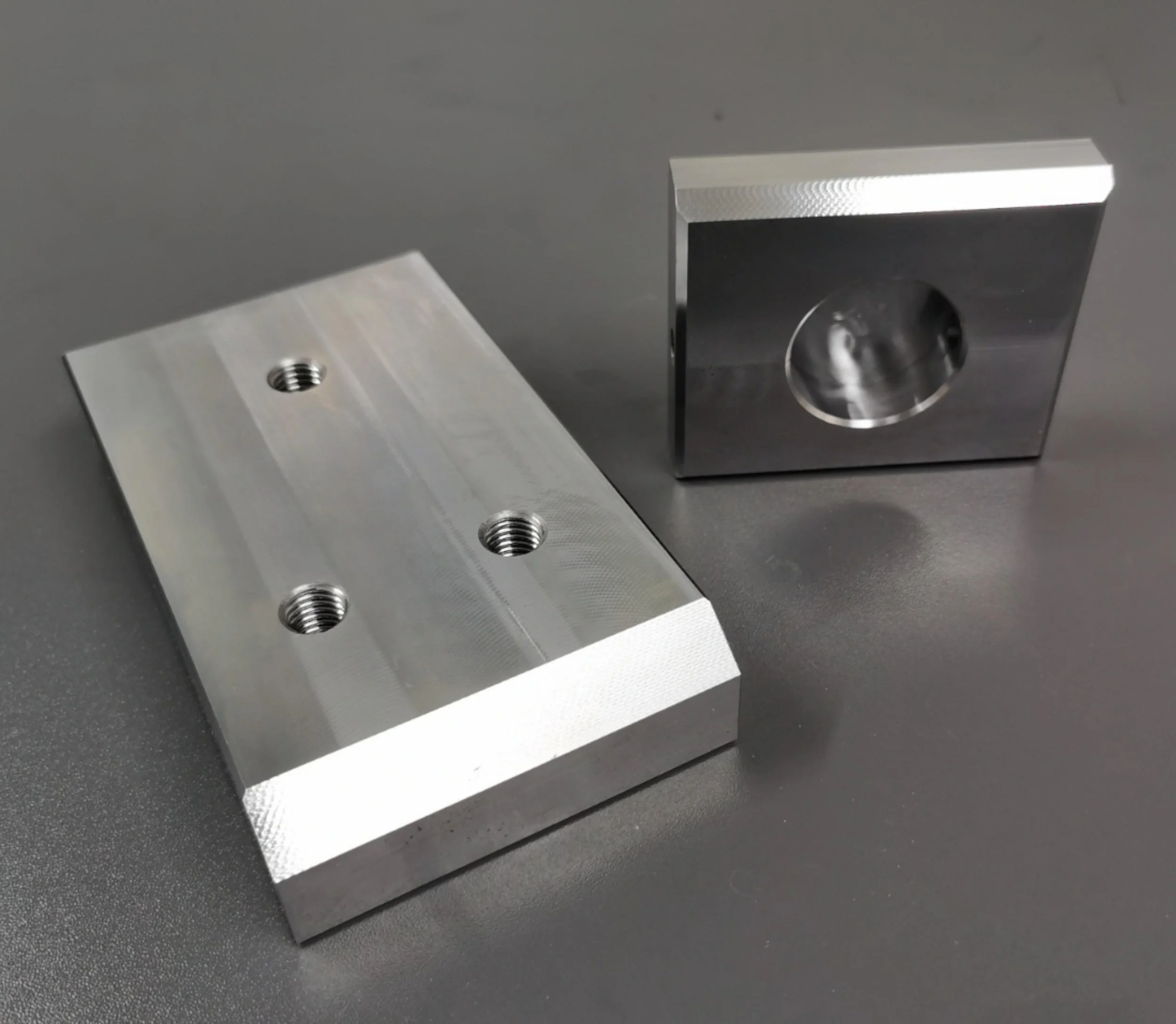 Two machined metal parts with threaded holes, one rectangular and one cube-shaped, placed on a dark surface.