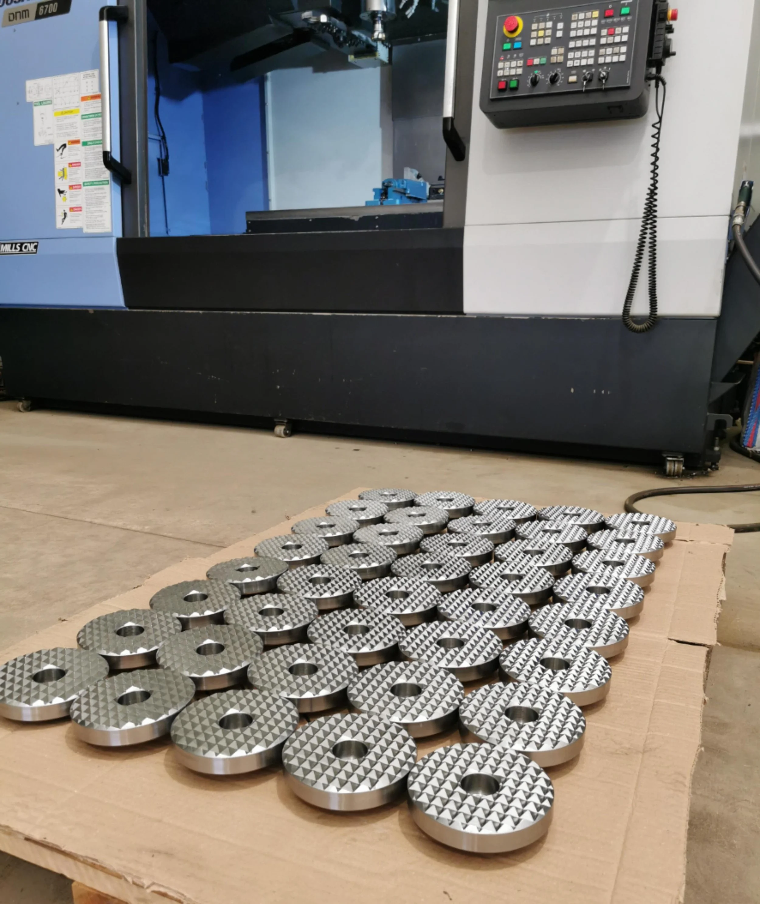 Metal industrial parts with textured surfaces arranged on a cardboard sheet in a workshop near a CNC machine.