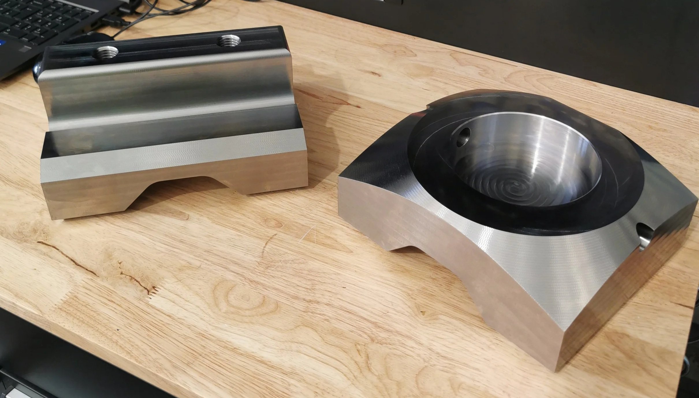 Two machined metal components with precise shapes and grooves on a wooden table.