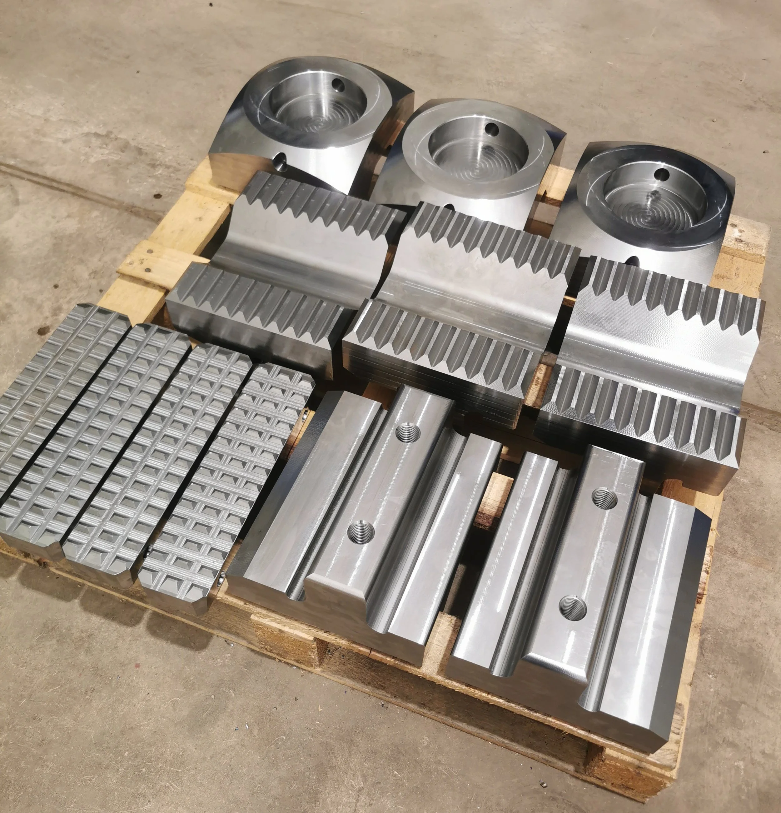 Set of machined metal parts with various grooves and holes, placed on a wooden pallet on a concrete floor.