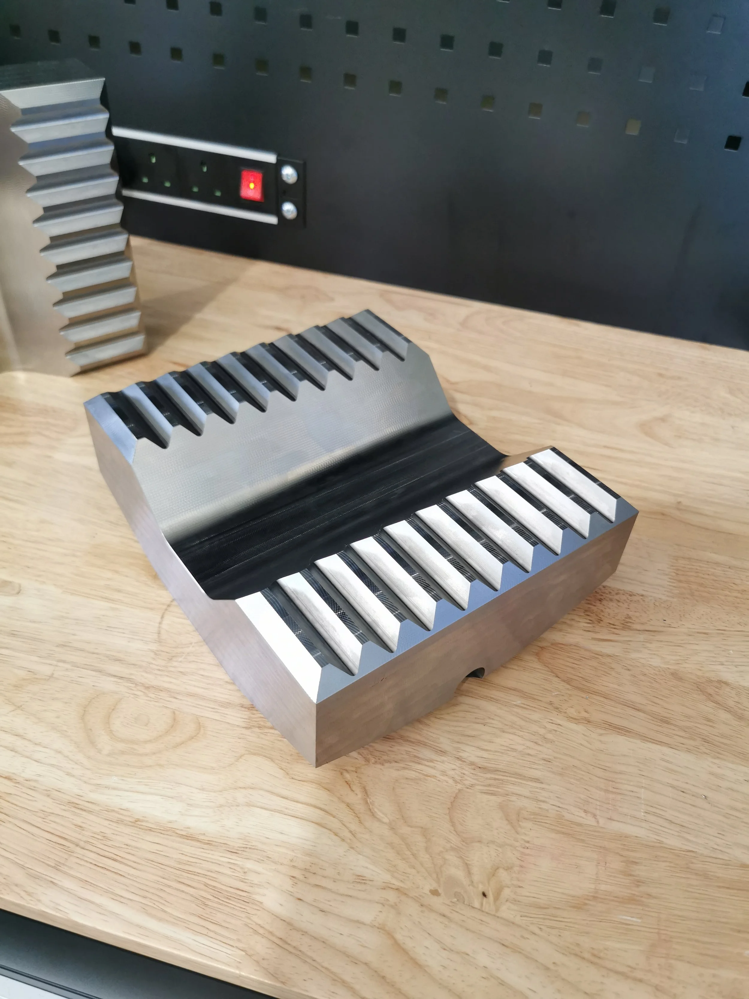 Metal mold with sections shaped like piano keys, placed on a wooden surface with a black pegboard and electrical outlet in the background.